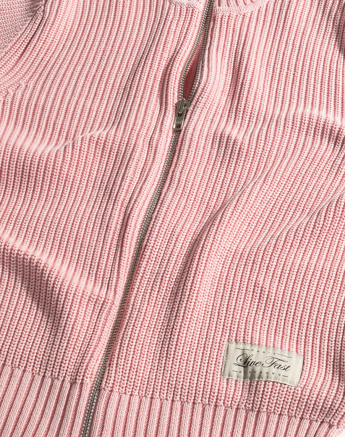 A close-up of the LFDY Knit Jacket by LIVE FAST DIE YOUNG in pink ribbed knit, featuring a zip-up front, Stehkragen, white "Live Fast" label at the hem, and made from 100% Baumwolle in a comfortable Regular Fit.