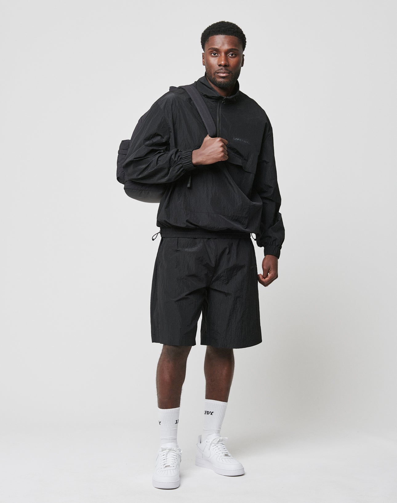 A man stands against a plain background in an oversized black windbreaker, LIVE FAST DIE YOUNG Live Fast Nylon Shorts, white socks and sneakers. He carries a black bag over one shoulder and looks straight at the camera.