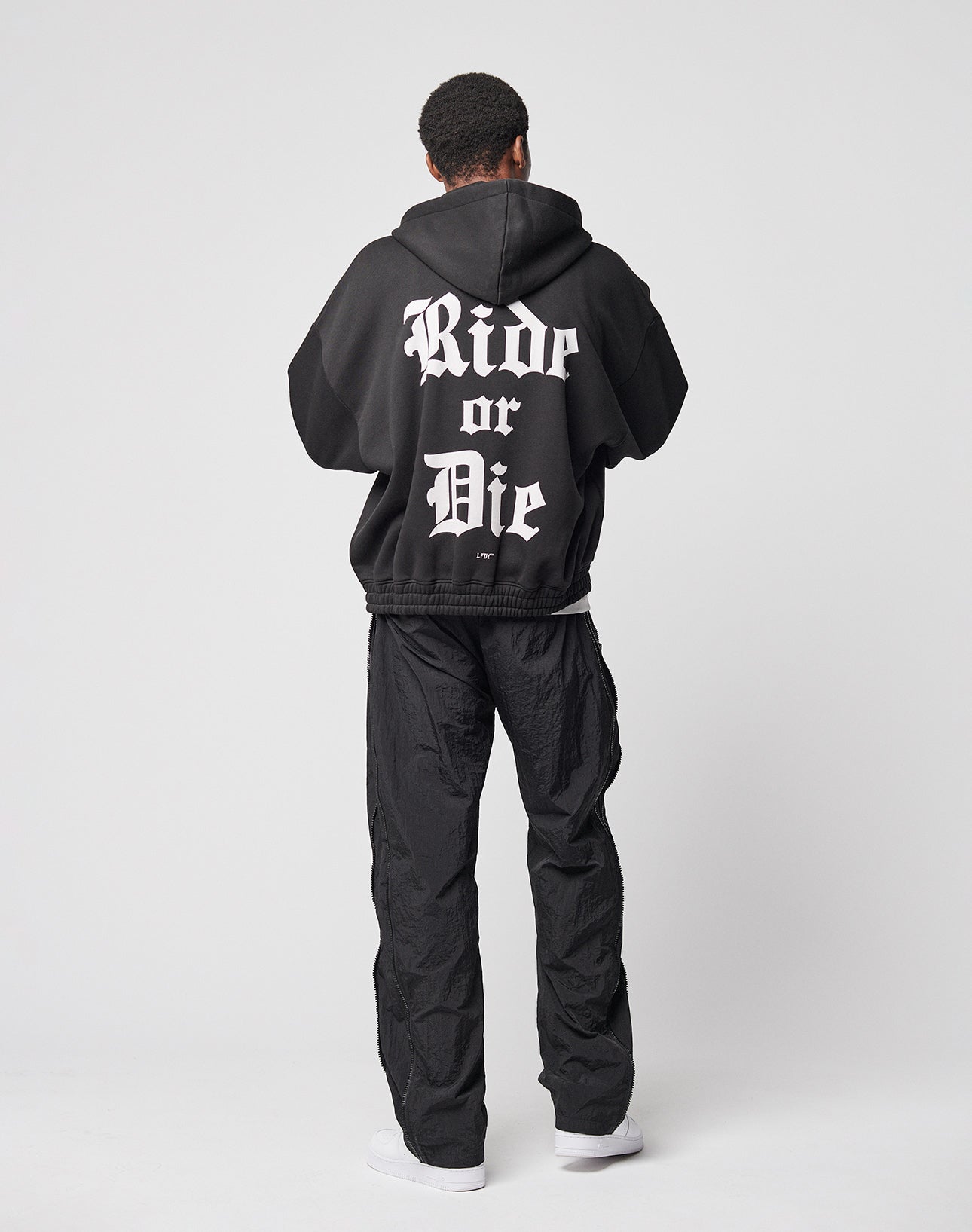 A person faces away, wearing the LIVE FAST DIE YOUNG Ride or Die Hooded Zip with doppellagige Kapuze and bold white “Ride or Die” gothic letters, paired with loose black pants (elastischer Bund) and white sneakers, against a white background.