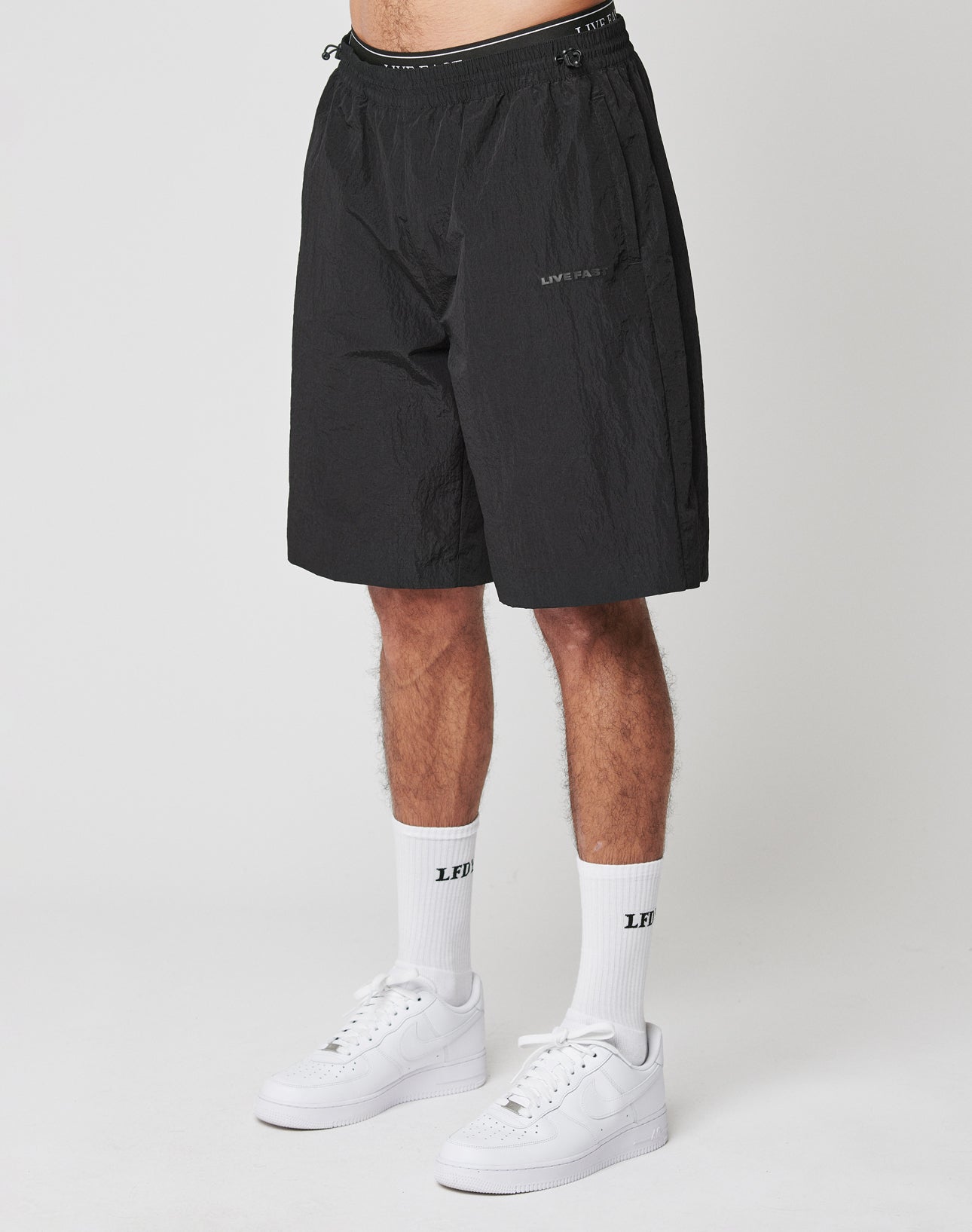 A person wears black Live Fast Nylon Shorts by LIVE FAST DIE YOUNG with a drawstring waist and oversized fit, paired with white crew socks featuring LFTS text and white sneakers, standing against a plain white background.