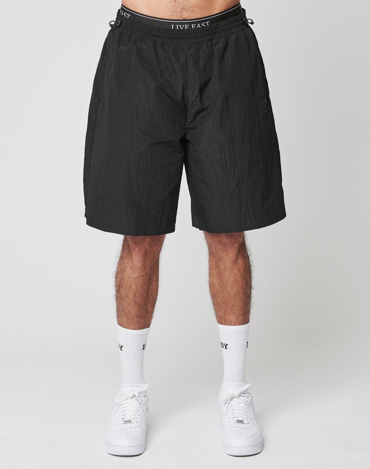 A person wears LIVE FAST DIE YOUNGs Live Fast Nylon Shorts, which feature an oversized fit and a bold Silikon-Print waistband, paired with white crew socks and sneakers. The setting is a plain light background; only the lower body is visible.