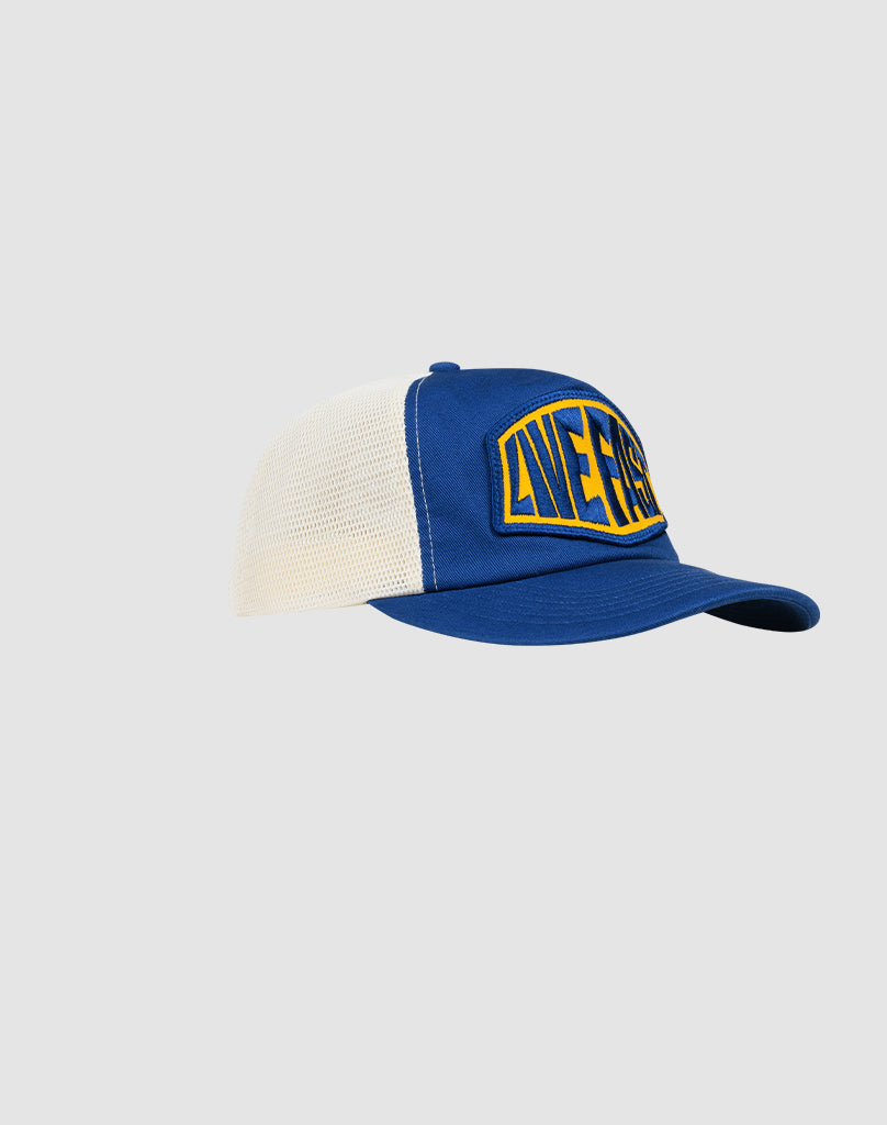The LFDY Patched Trucker by LIVE FAST DIE YOUNG is a blue and white trucker cap with snap closure, curved brim, and a yellow-blue LIFERS logo patch on the front, shown against a plain light gray background.
