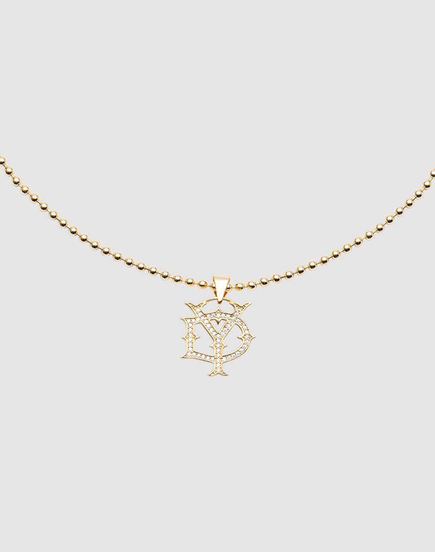 The DY Chain by LIVE FAST DIE YOUNG is a gold-plated beaded necklace with a cubic zirconia pendant featuring stylized overlapping D and Y letters, shown on a plain light gray background.