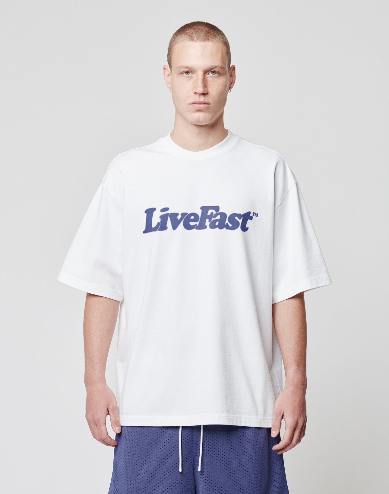 A person stands facing forward, wearing a LIVE FAST DIE YOUNG Basic Tee (100% cotton, white) with LiveFast in bold dark letters, paired with blue athletic shorts against a plain light background.