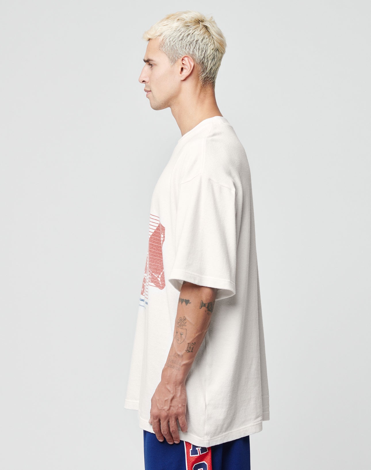 A man with short blond hair and tattoos on his left arm stands in profile wearing the LIVE FAST DIE YOUNG LFDY Juveniles Tee, an oversize vintage white 100% Baumwolle shirt with a red and blue graphic, paired with blue shorts, against a light gray background.