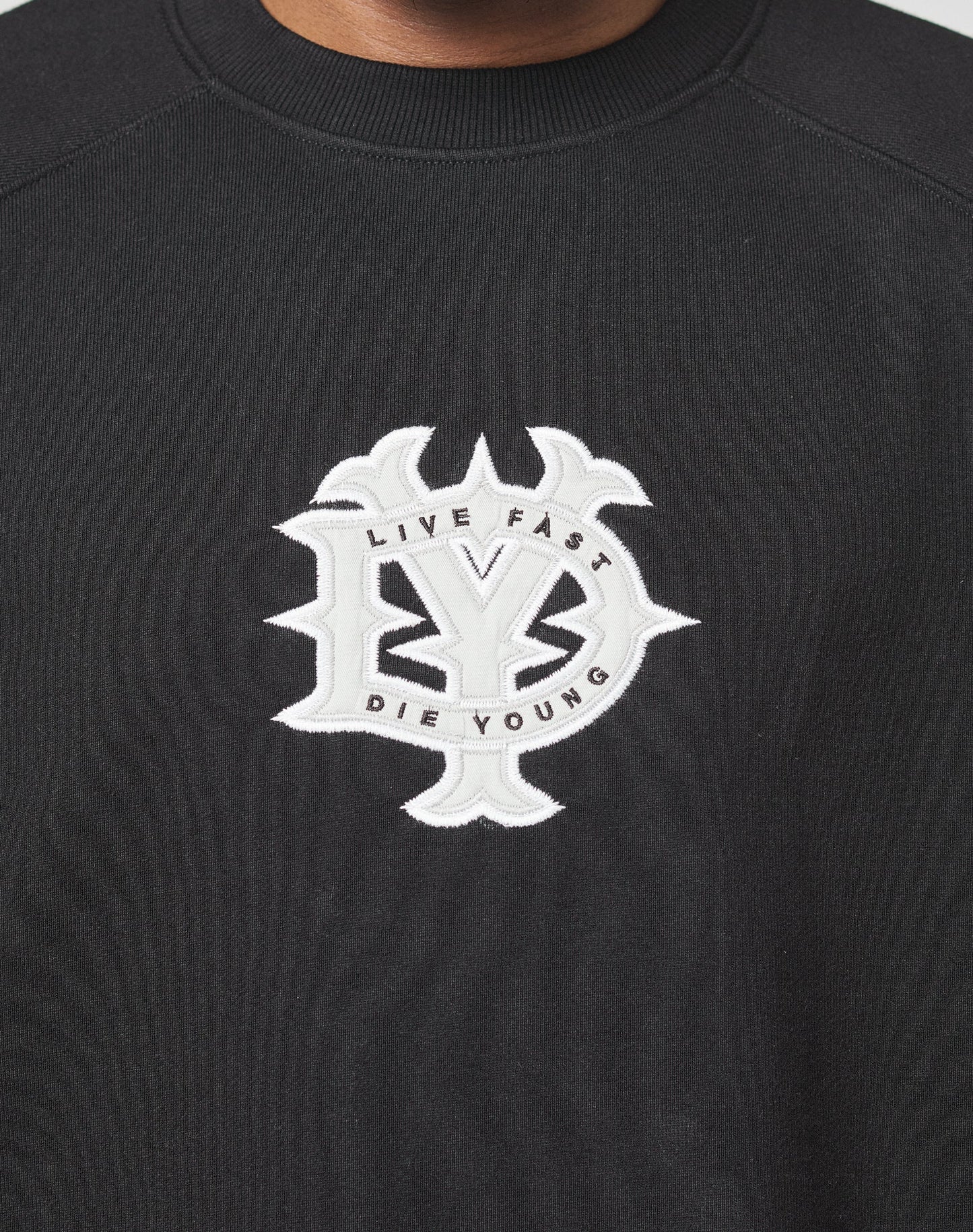 The DY Sweater by LIVE FAST DIE YOUNG is a black oversized sweatshirt with raglan sleeves, featuring a large white embroidered LY emblem and LIVE FAST DIE YOUNG in a circular chest design.