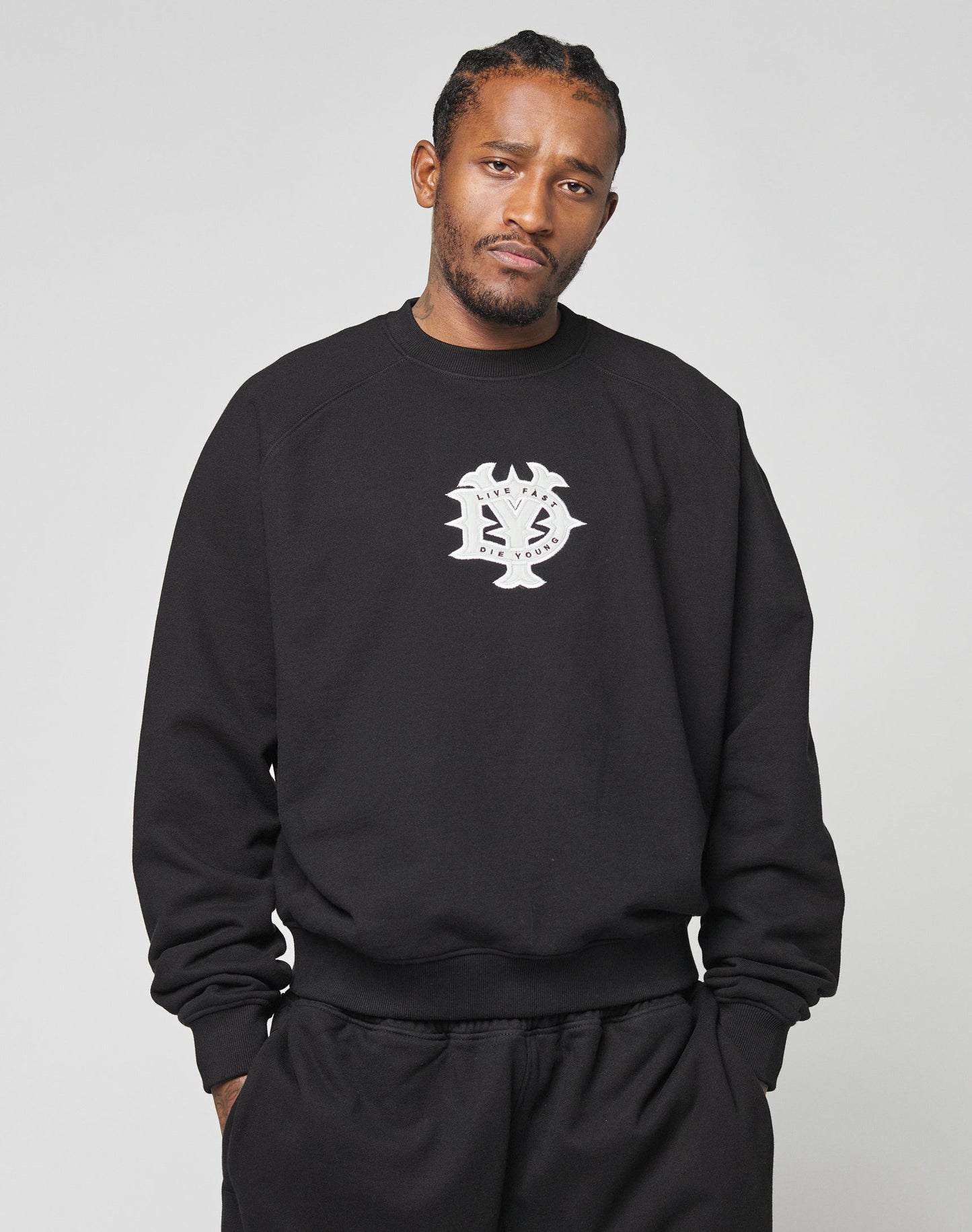 A man with braided hair stands against a light gray background, wearing the black DY Sweater by LIVE FAST DIE YOUNG, featuring Raglan sleeves and a white logo on the chest, paired with matching black pants.