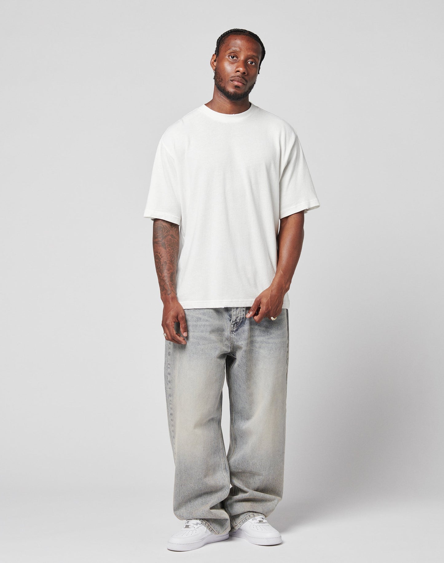 A man with short hair and tattoos on his left arm stands against a plain background, wearing an oversize fit LIVE FAST DIE YOUNG LFDY Vintage Tee in 100% Baumwolle, paired with baggy light-wash jeans and white sneakers. He has a neutral expression.