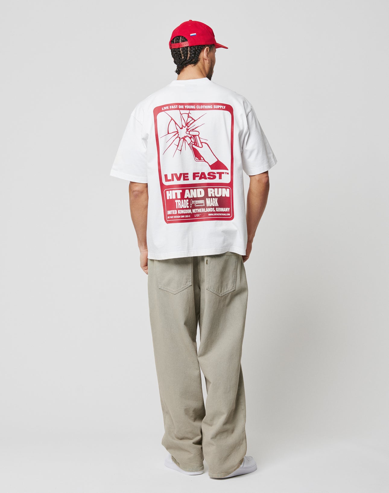A person in an oversized white Hit and Run Tee by LIVE FAST DIE YOUNG, with bold red LIVE FAST text on the back, 100% Baumwolle, beige pants, and a red cap stands facing away against a plain light gray background.