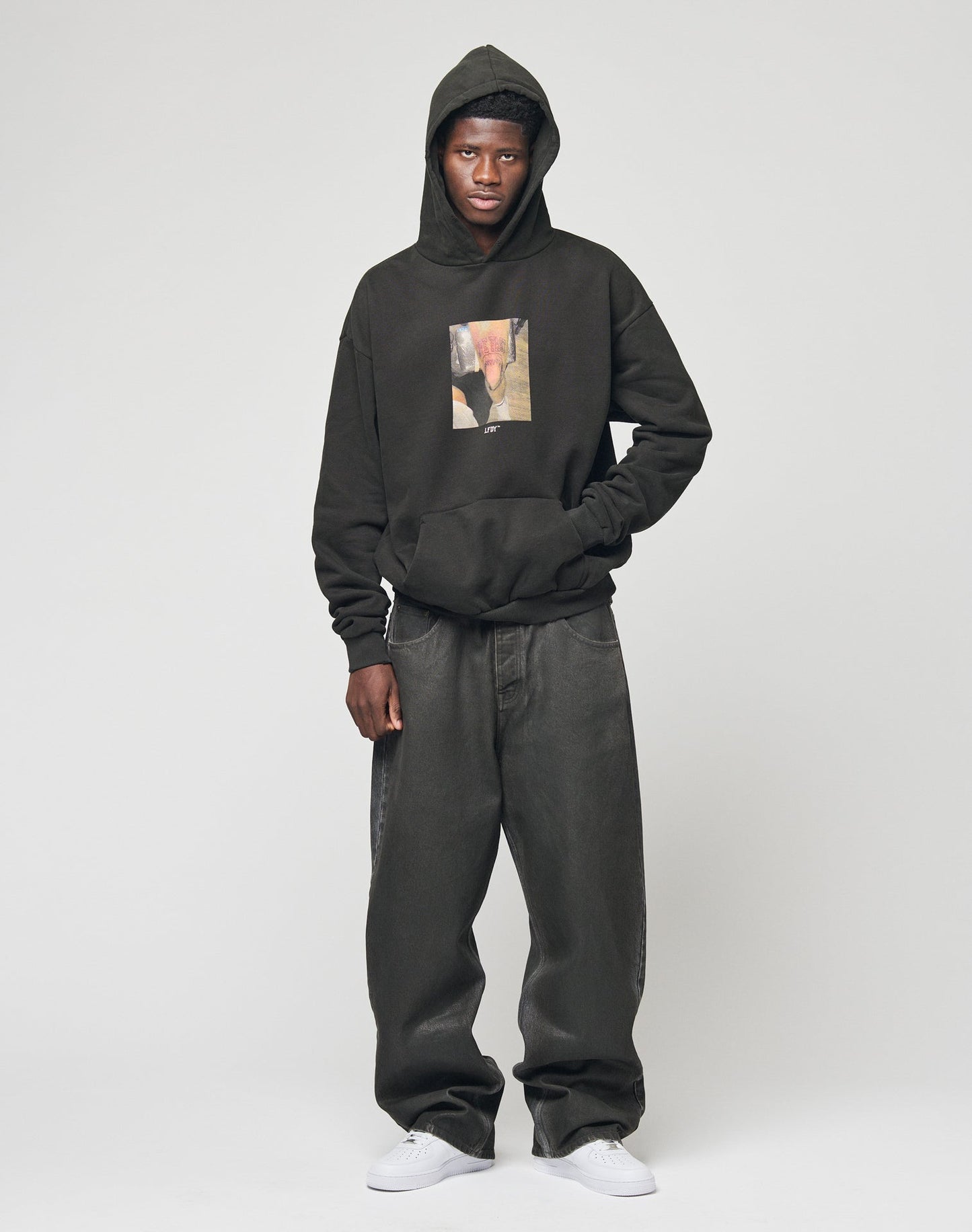 A person stands against a plain background wearing a black hoodie with a photo print, LFDY Coated Baggy Denim jeans by LIVE FAST DIE YOUNG, and white sneakers. The hood is up and their hands are by their sides.