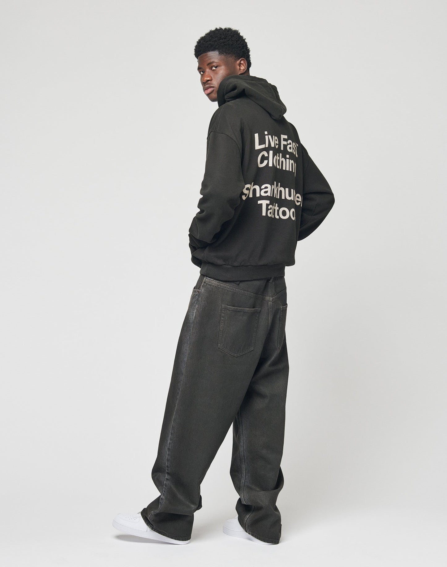 A man faces away, wearing a black LFDY Sharkhunter Hooded sweatshirt by LIVE FAST DIE YOUNG with white “Live Fast Clothing, Sharkhuge Tattoo” text on the back, loose dark jeans, and white sneakers.