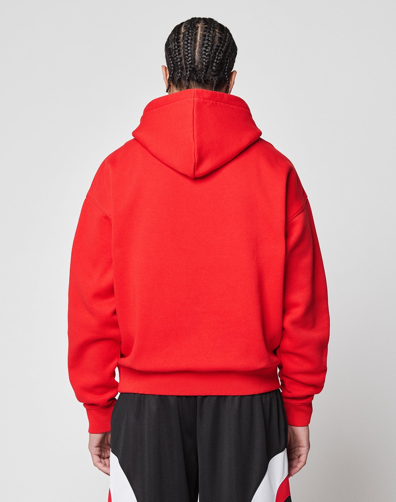 Seen from behind, a person with braided hair wears the LIVE FAST DIE YOUNG Live Fast College Hooded sweatshirt in bright red, styled oversized, paired with black pants featuring red and white accents against a plain light background.