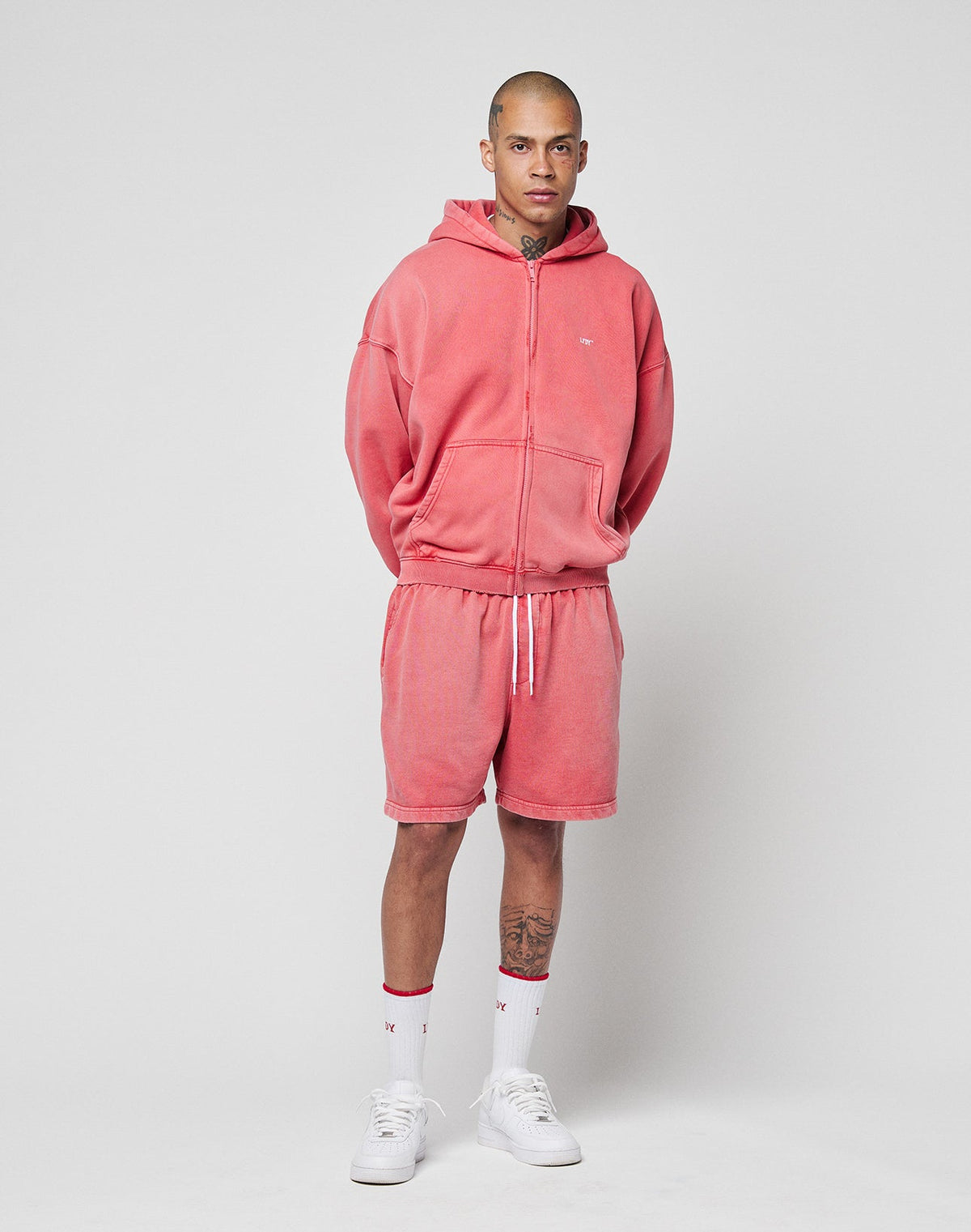 A person stands against a plain background wearing the LIVE FAST DIE YOUNG Basic Hooded Zip and matching shorts with individuellerm Sprayeffekt, paired with white socks and sneakers. Their hands are behind their back, tattoos visible on neck and legs.