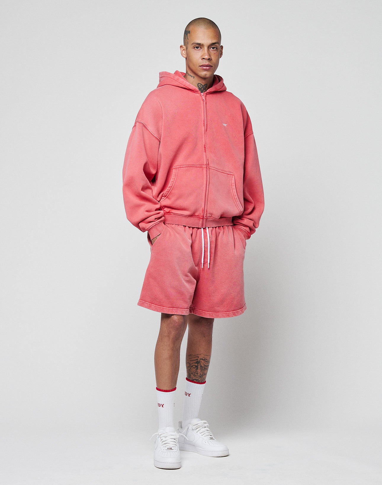 A person stands before a plain light background wearing LIVE FAST DIE YOUNG’s Basic Sweat Shorts and matching pink hoodie with elastic waistband, white crew socks with red text, and white sneakers. The hoodie is zipped up as they look at the camera.