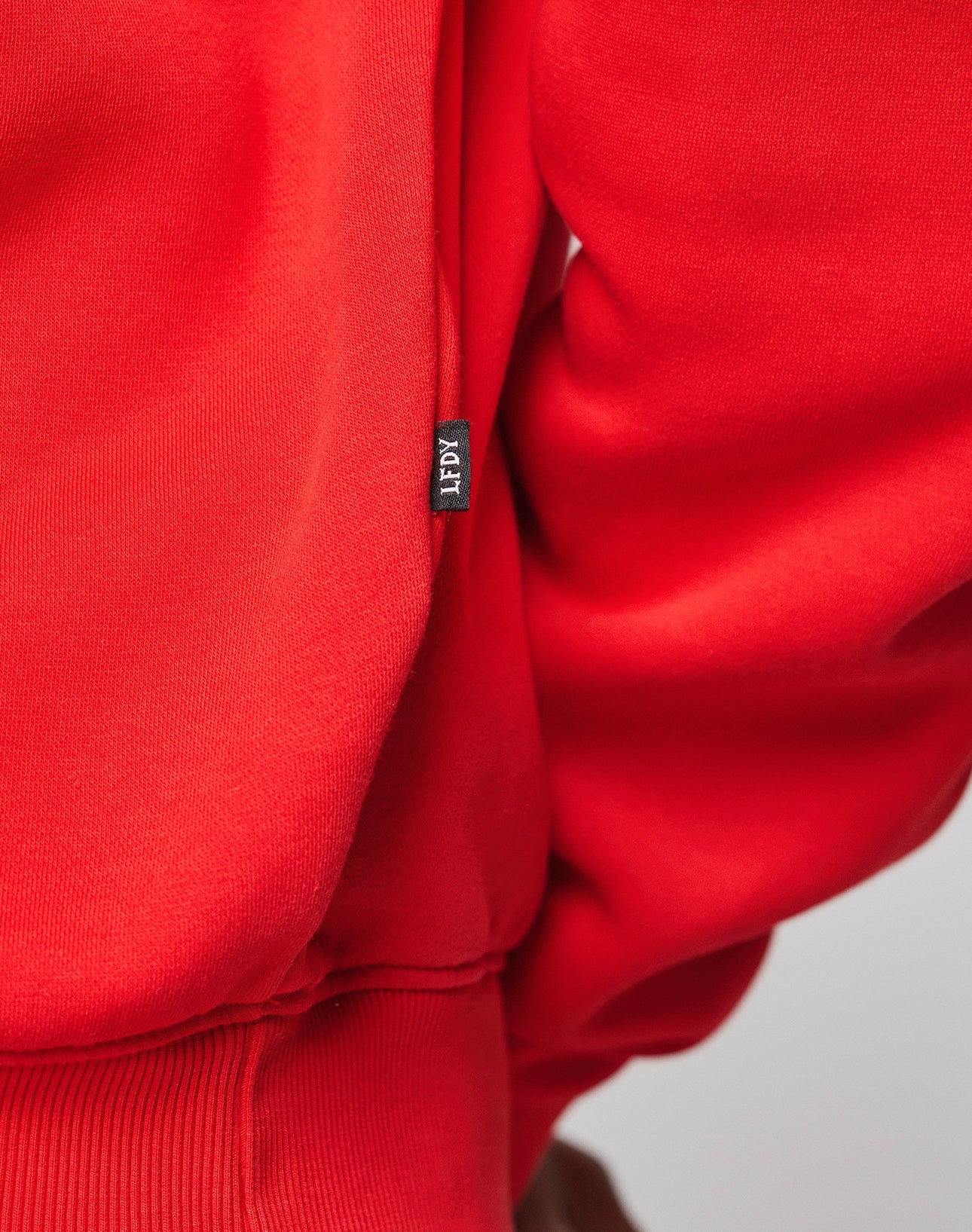 Close-up of a bright red Live Fast College Hooded sweatshirt by LIVE FAST DIE YOUNG, featuring a small black fabric tag with the brand name in white attached at the side seam.