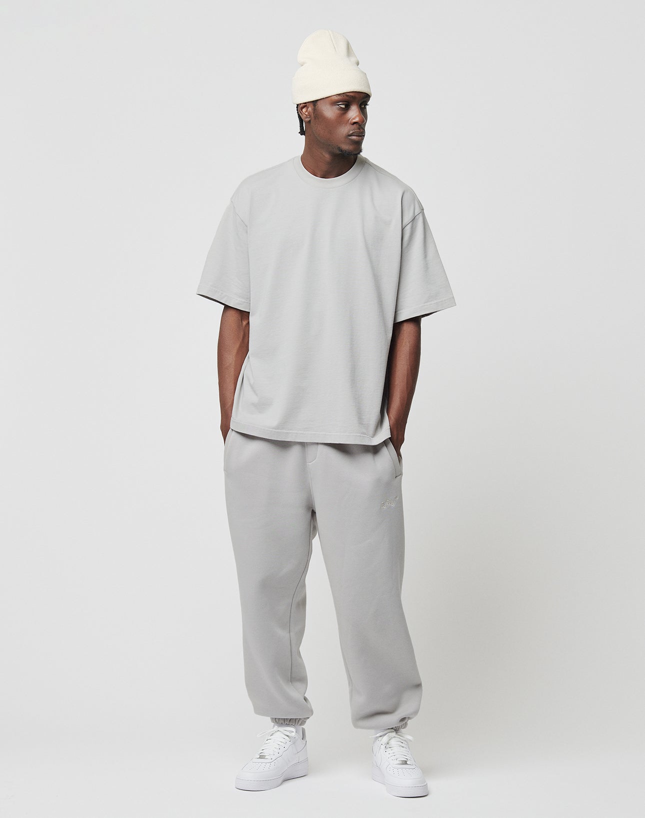 A person stands against a white background, wearing the LIVE FAST DIE YOUNG Basic Heavy Tee in oversize fit light gray 100% Baumwolle, with matching sweatpants, white sneakers, and a cream beanie. They look to the side with hands in pockets.