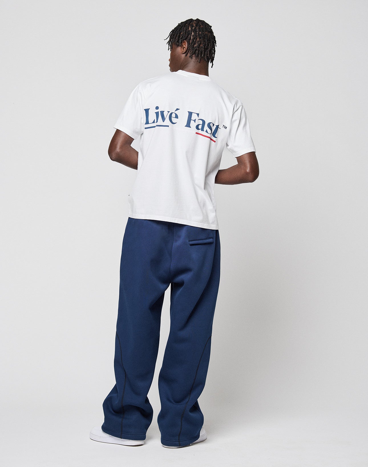 A person stands facing away, showcasing the LIVE FAST DIE YOUNG Live Fast Paris Tee, a regular fit white t-shirt with blue and red Live Fast front and back print, paired with loose navy pants and white shoes against a plain white background.