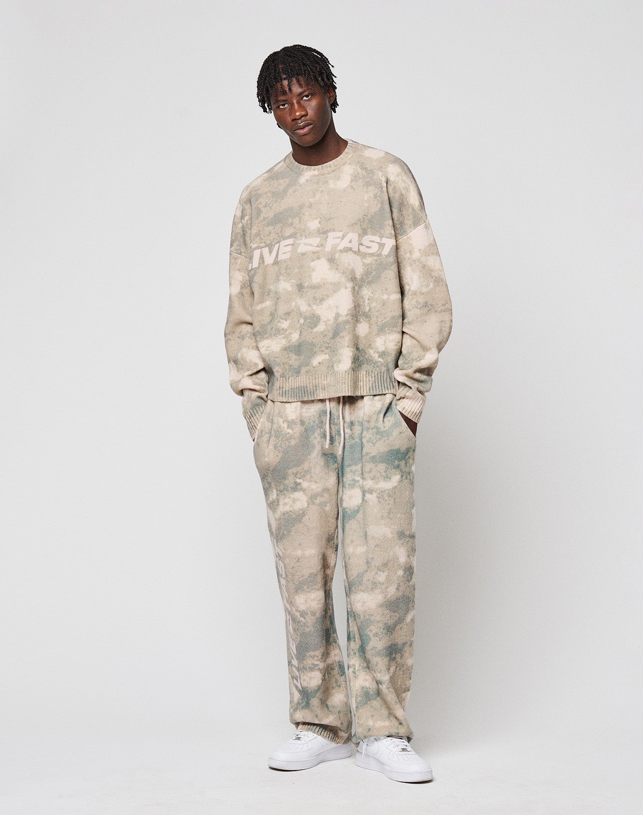 Wearing the LIVE FAST DIE YOUNG Live Fast Boucle Sweater in an oversize beige and green tie-dye fit, with matching pants and white sneakers. The set features the signature Live Fast Stick and is made from soft polyester.