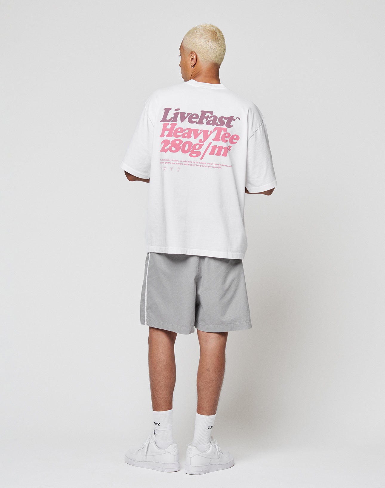 A person with short blond hair faces away, wearing the LIVE FAST DIE YOUNG Basic 280 Tee in oversize fit with bold pink backprint, paired with light gray shorts, white socks, and white sneakers against a plain white background.