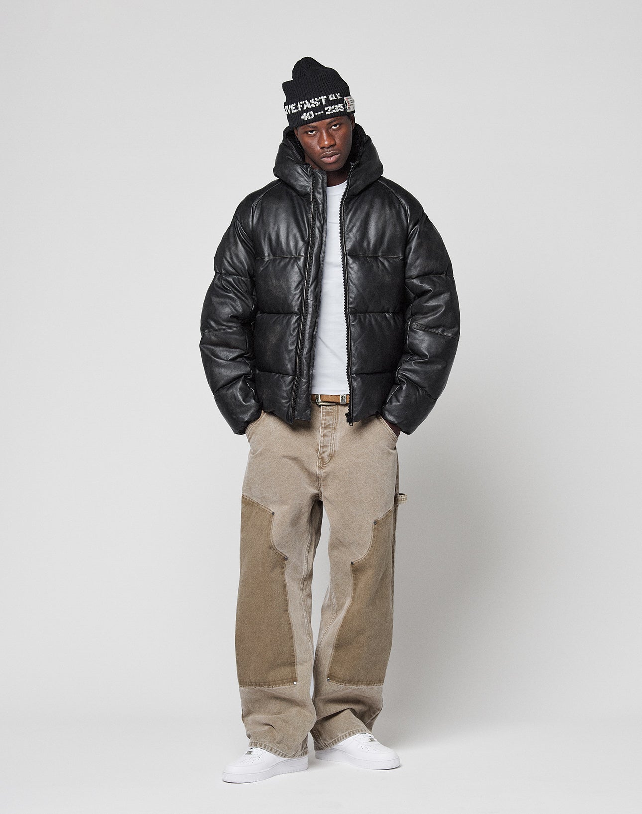 A person stands against a plain background wearing the LIVE FAST DIE YOUNG Live Fast Leather Puffer Jacket, beige wide-leg patchwork jeans, a white t-shirt, white sneakers, and a black beanie with white text.