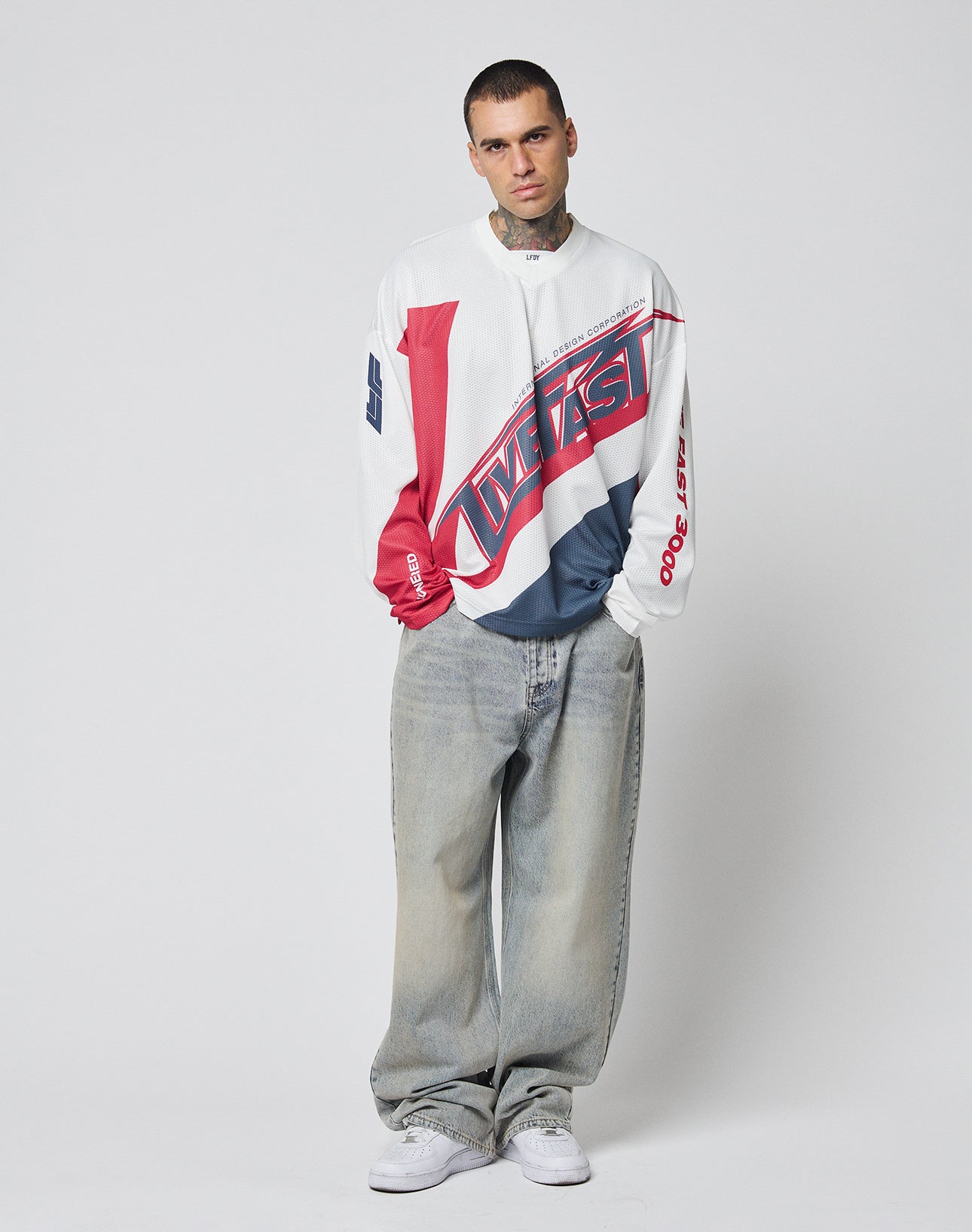 A man stands against a white background wearing the LIVE FAST DIE YOUNG LFDY Racetrack Jersey—an oversize, color-blocked long-sleeve shirt with red, white, and navy multi-logo prints—baggy light-wash jeans, and white sneakers. He looks at the camera neutrally.