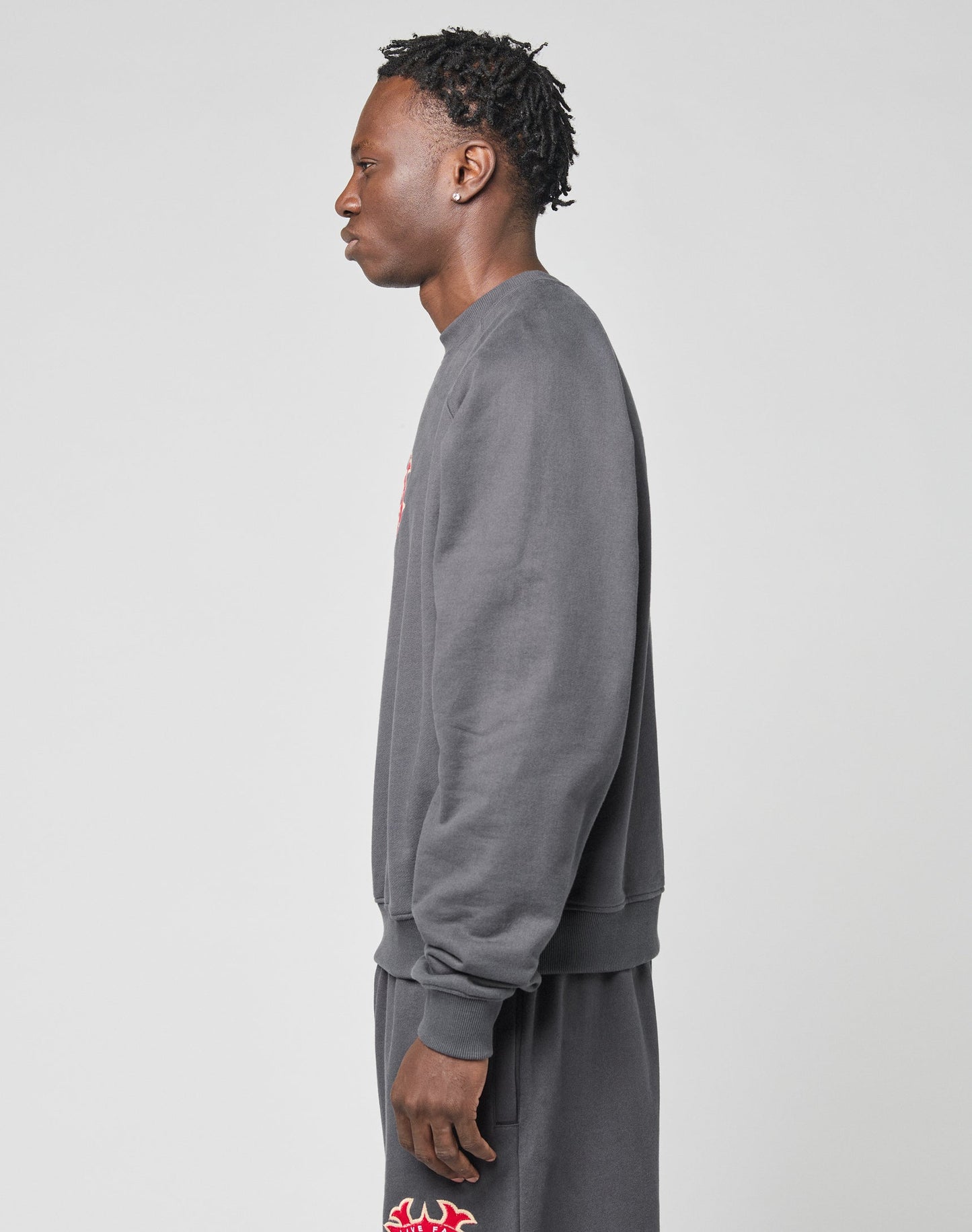 A person stands in profile against a light gray background, wearing the LIVE FAST DIE YOUNG DY Sweater in an oversized dark gray fit with raglan sleeves, paired with matching shorts featuring red and white graphic details.