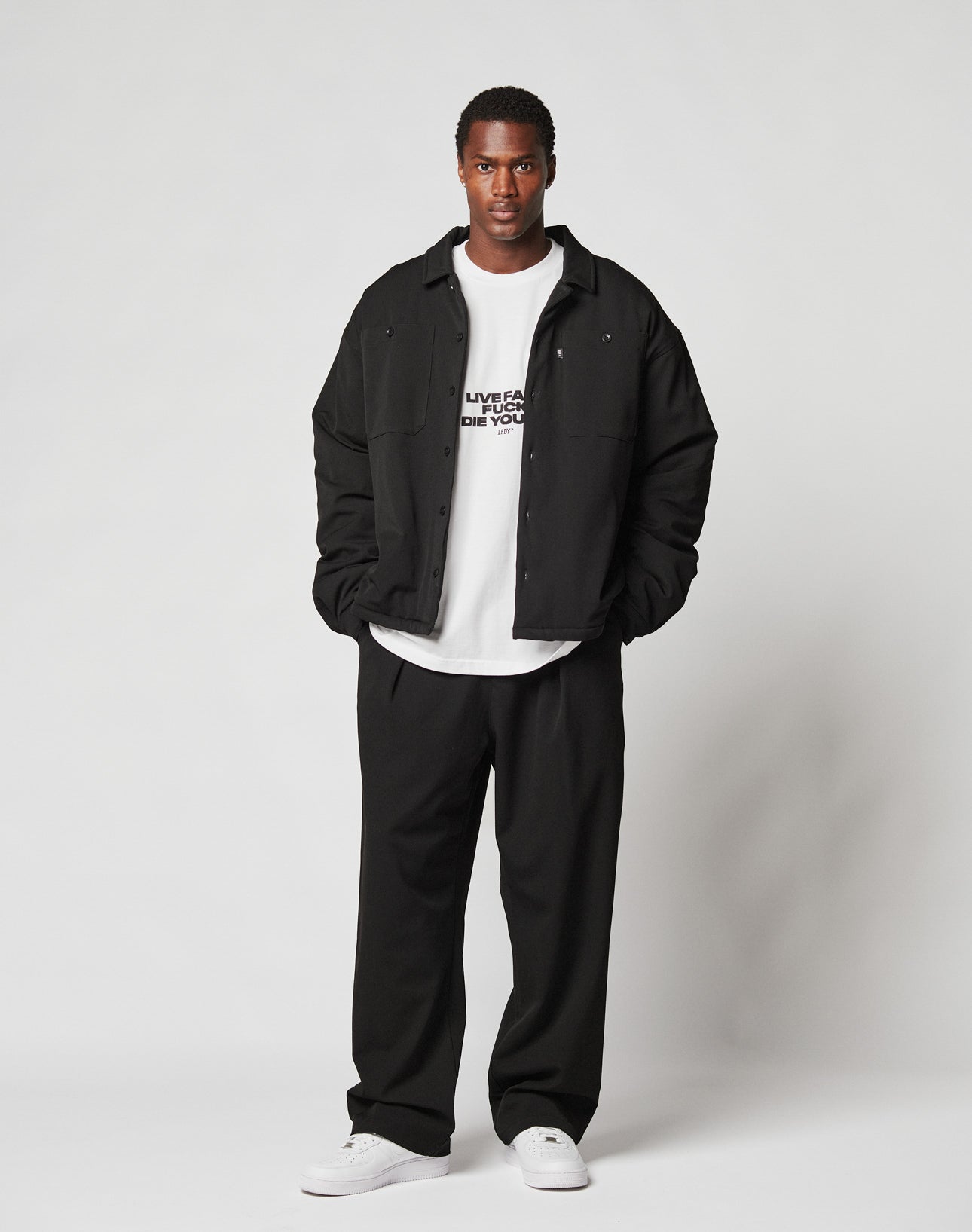 A man faces forward in a black Oversize Fit jacket over the WHITE Fuck Die Young Tee by LIVE FAST DIE YOUNG, loose black pants, and white sneakers. The background is plain and light-colored.