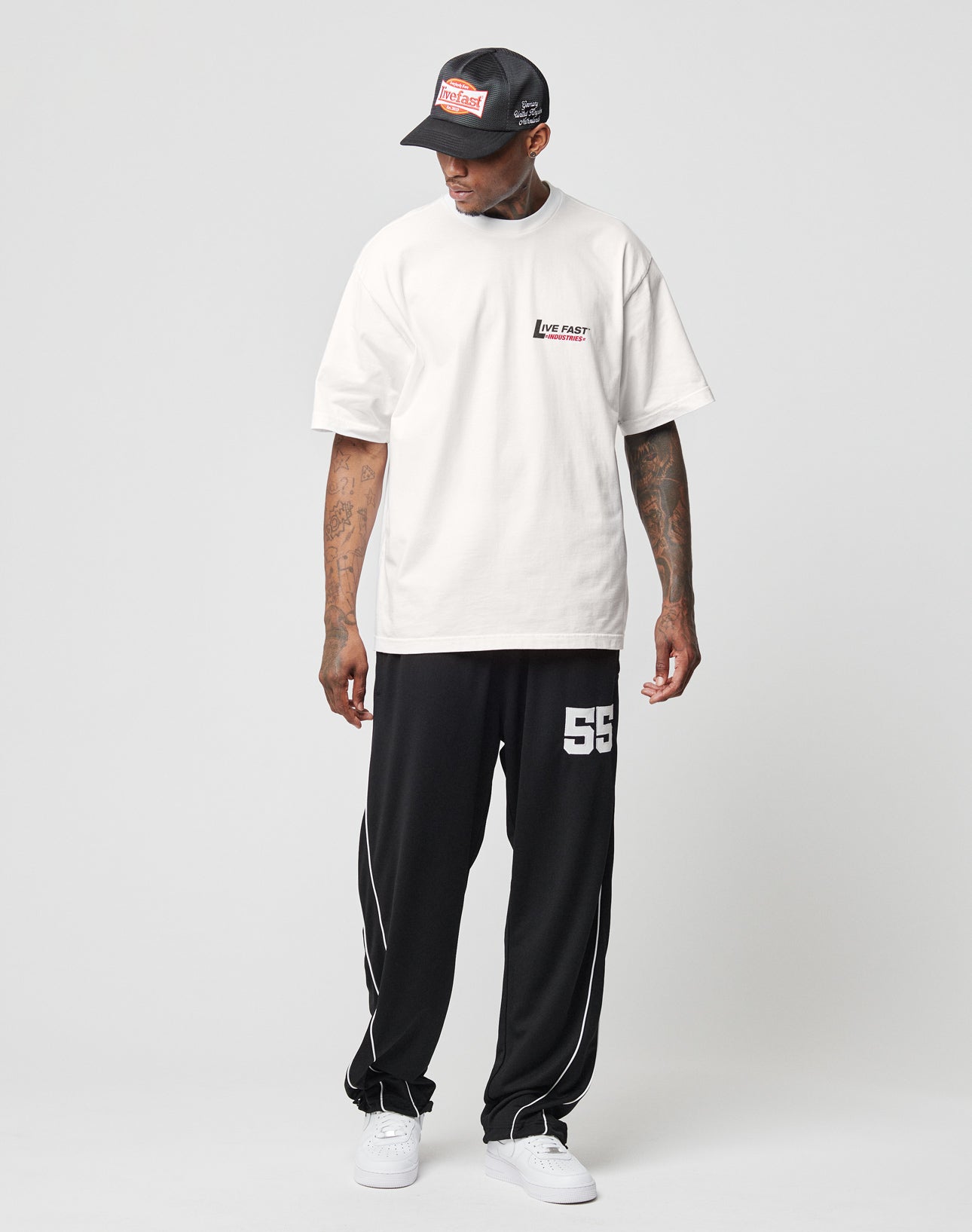 Man wearing an LFDY Industries Tee by LIVE FAST DIE YOUNG, a black cap, loose black pants with white 55 on the left thigh, and white sneakers, standing and looking to his left against a plain background.