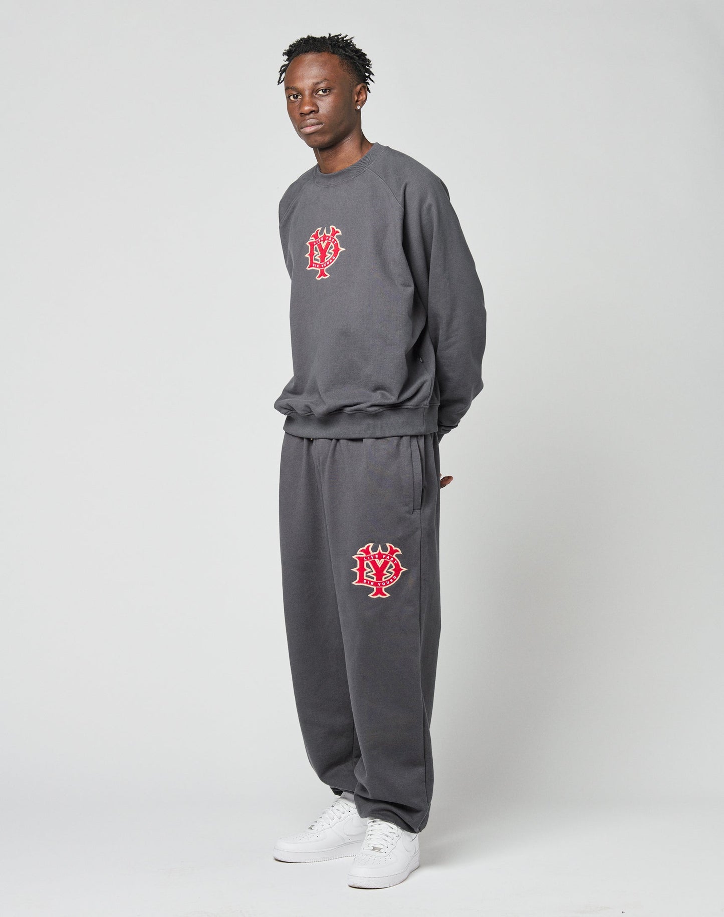 A person stands against a plain background wearing the LIVE FAST DIE YOUNG DY Sweater, an oversize fit dark gray sweatshirt and sweatpants set with raglan sleeves and red and white “NY” logos, paired with white sneakers.
