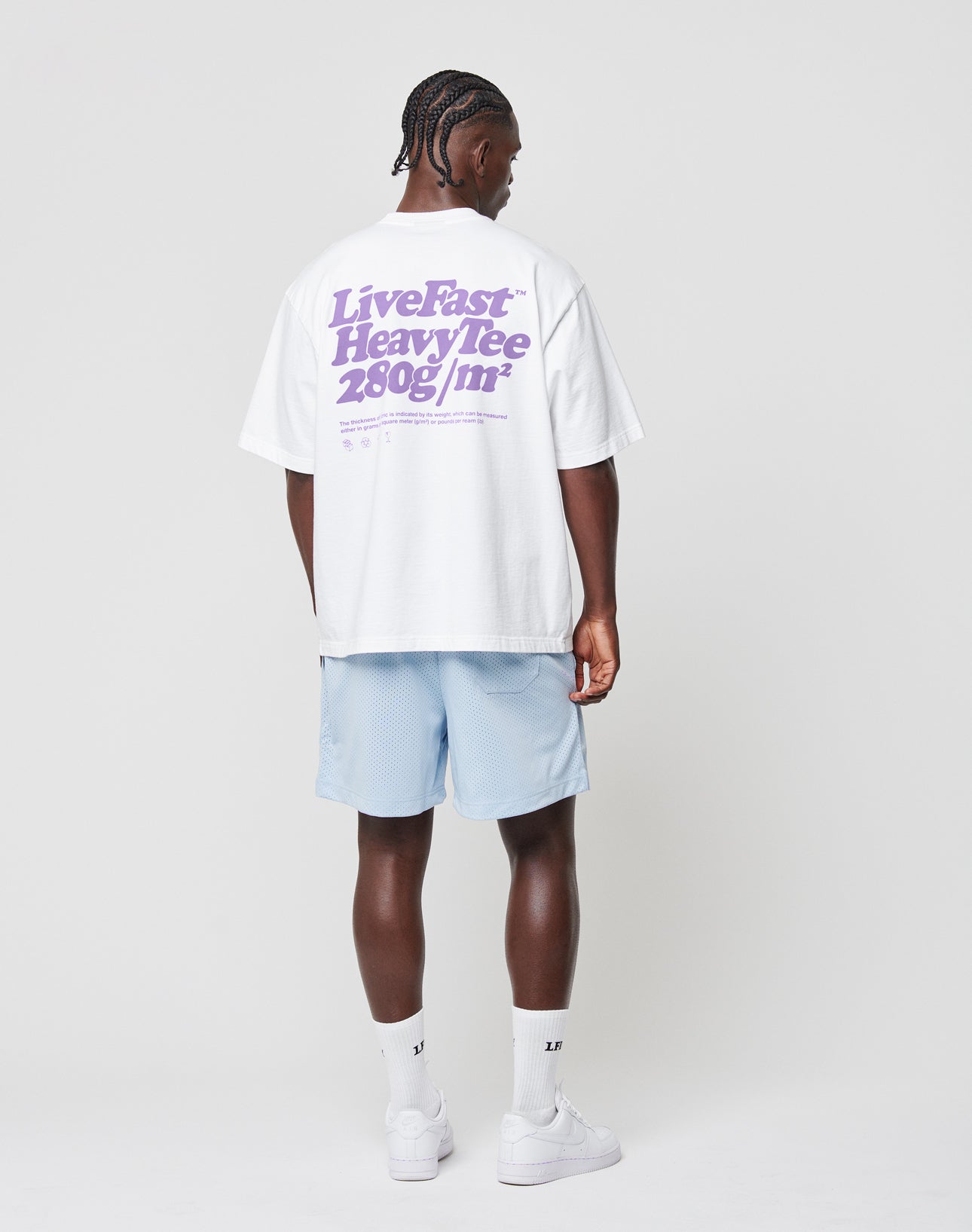 A person faces away, wearing a white LIVE FAST DIE YOUNG Basic 280 Tee with purple text, light blue shorts, white socks, and sneakers against a plain white background. The Oversize Fit tee is made from 100% Baumwolle (cotton).