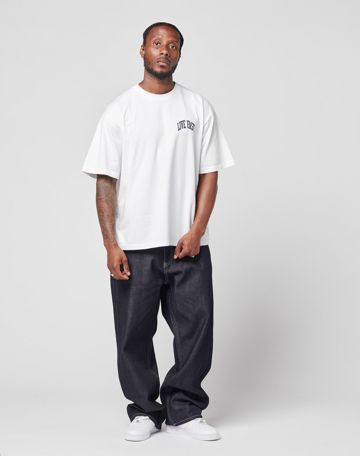 A man stands against a plain background wearing the LIVE FAST DIE YOUNG Live Fast London Tee, featuring small front and back prints, along with baggy dark jeans and white sneakers. His expression is neutral and his arms rest by his sides.