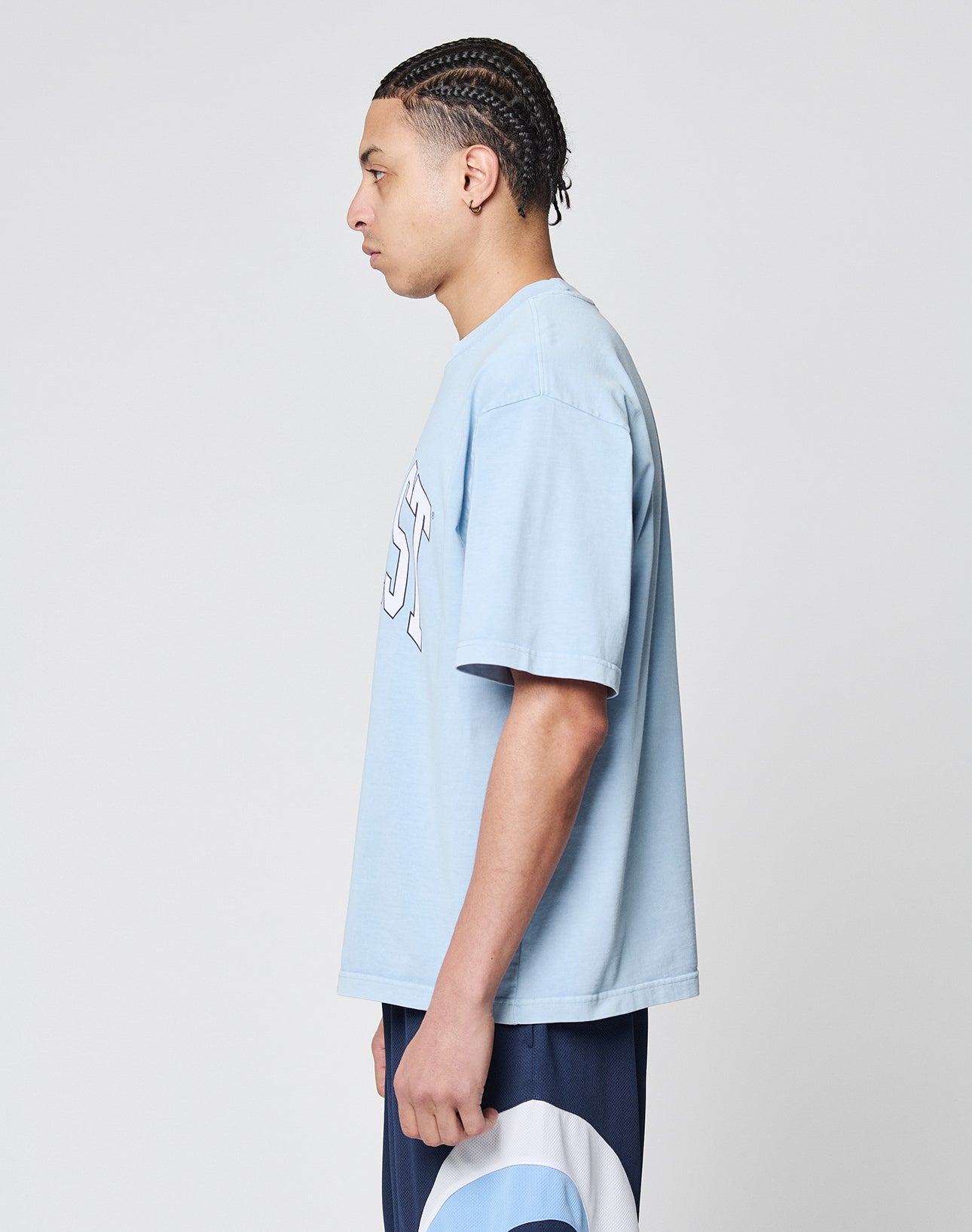A young man with braided hair stands in profile, wearing the LIVE FAST DIE YOUNG Live Fast College Tee in light blue with white lettering and navy and light blue shorts, set against a plain light gray background.