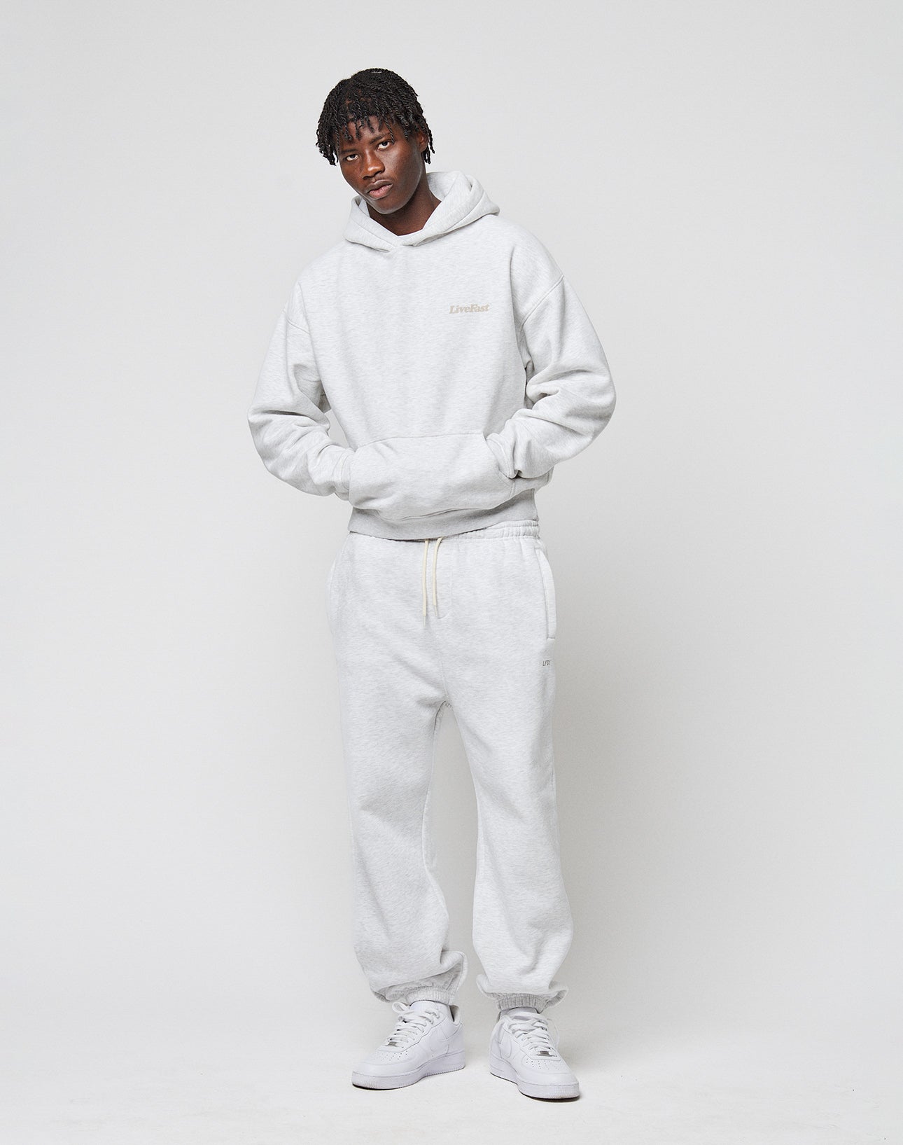 A young man stands against a white background, wearing the LIVE FAST DIE YOUNG Basic 360 Hooded in light gray with matching sweatpants and white sneakers. He has short, styled hair and his hands are clasped in front.