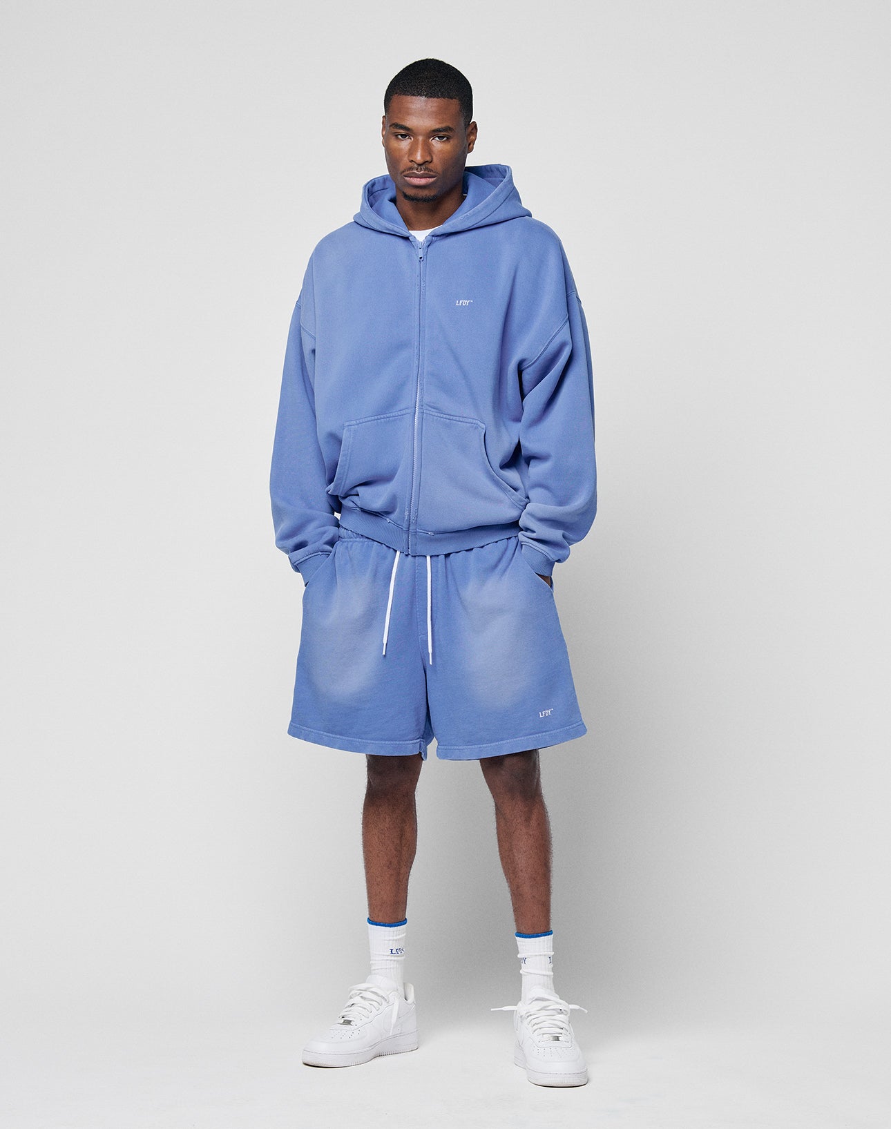 A man stands against a plain light background wearing the LIVE FAST DIE YOUNG Basic Hooded Zip with handgefertigtem Fade-Effekt and matching blue shorts, styled with white socks, white sneakers, hands in his pockets, looking at the camera.