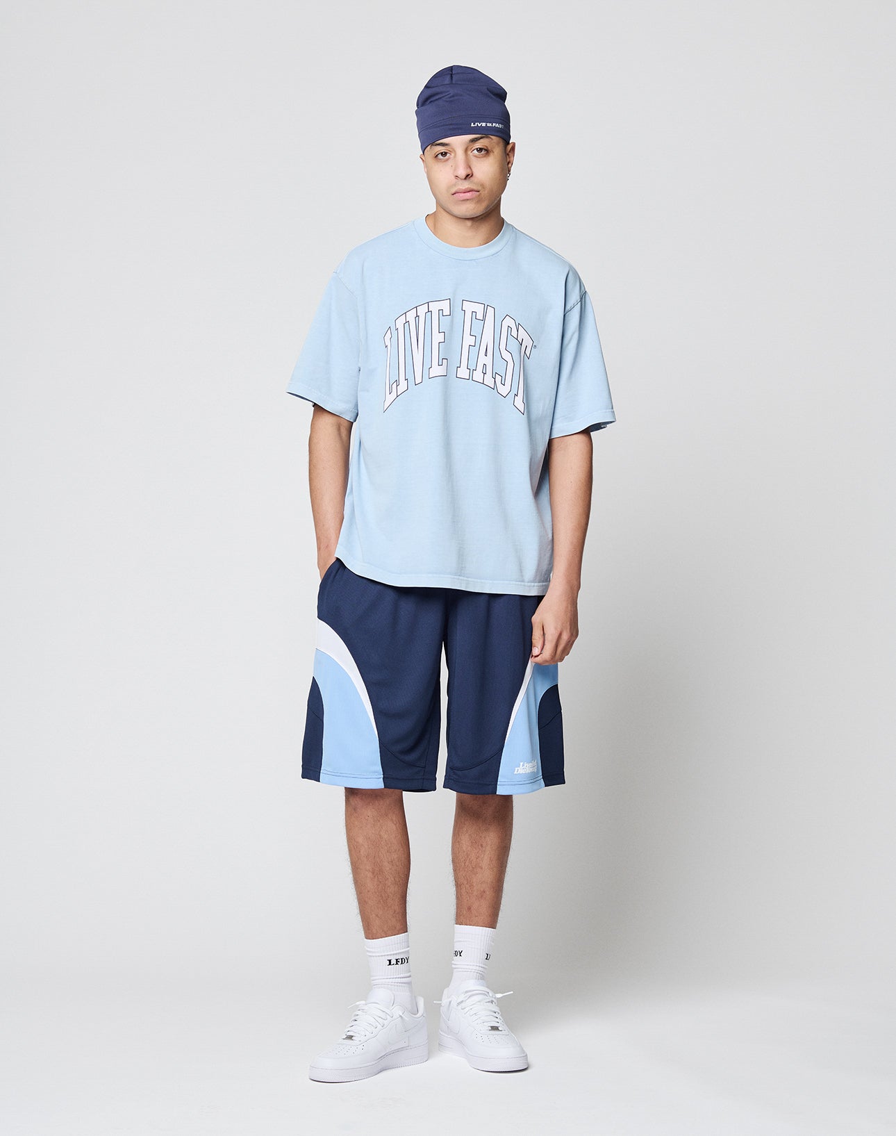A person stands against a plain background wearing a blue headwrap, LIVE FAST DIE YOUNG’s Live Fast College Tee in light blue with LIVE FAST printed on it, dark blue athletic shorts with light blue accents, white socks, and white sneakers.