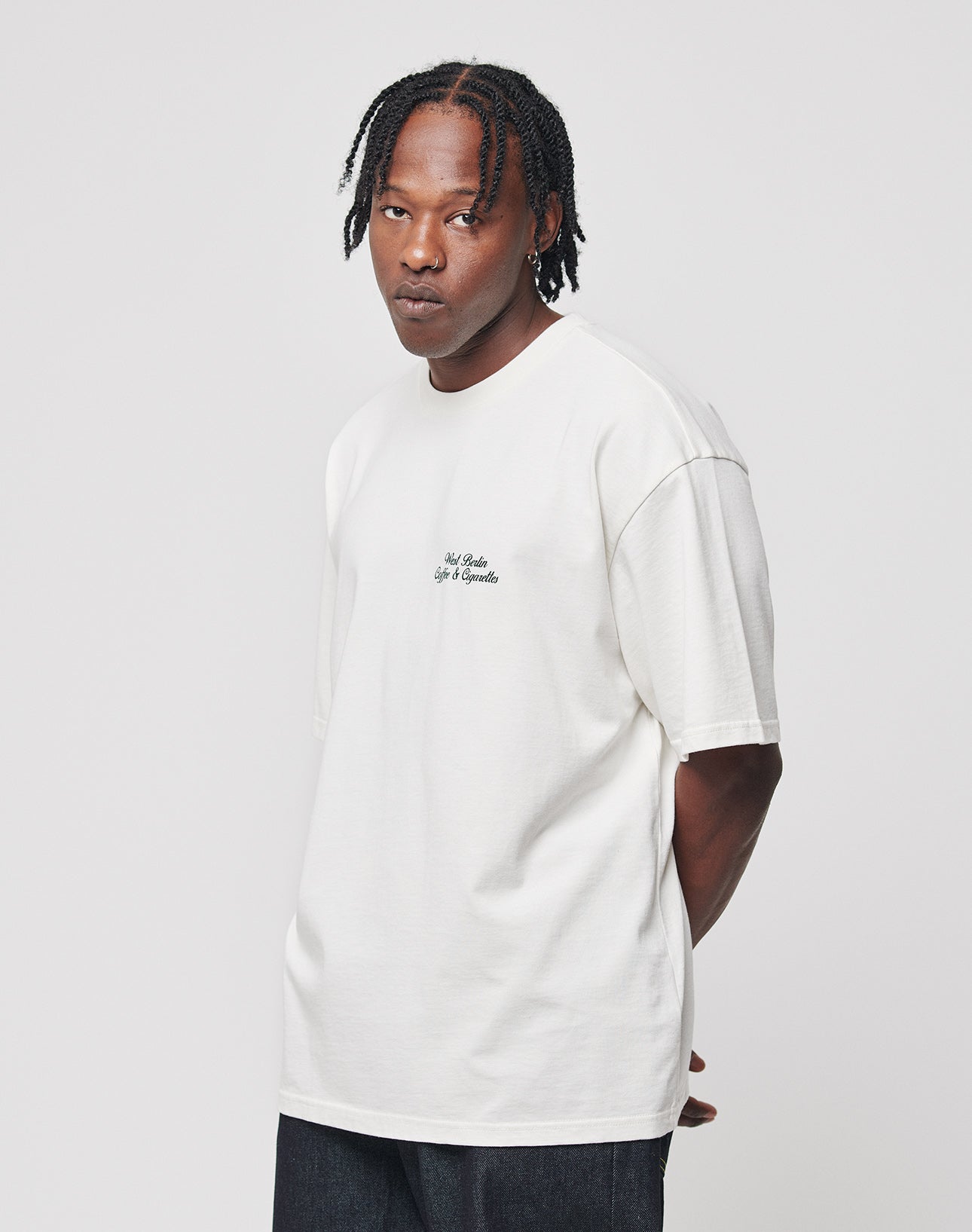 A man with short, braided hair wears the LIVE FAST DIE YOUNG Live Fast Coffee Tee— a loose white 100% cotton T-shirt with small chest text— and dark jeans, standing against a plain white background with his hands behind his back.