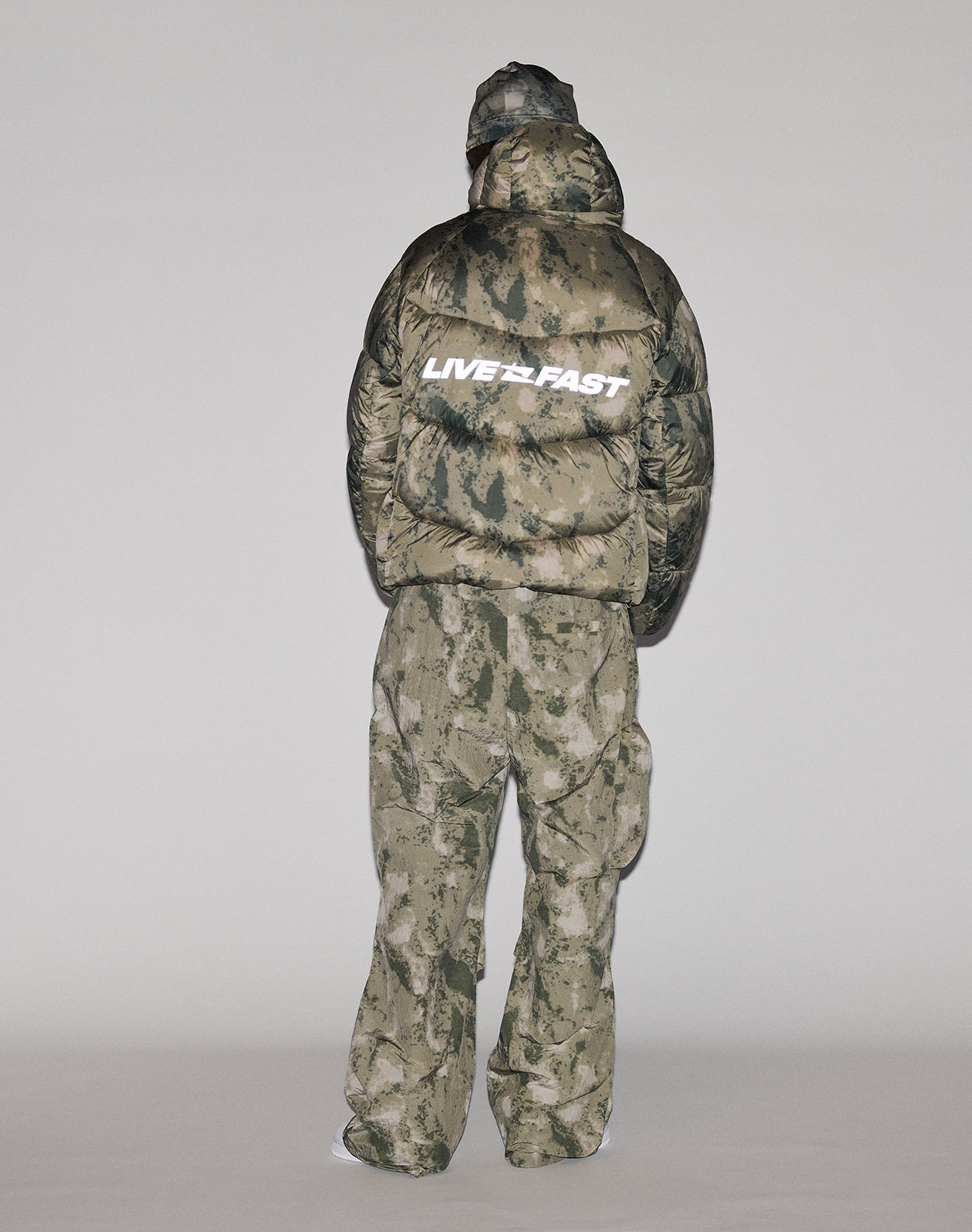 A person faces away, wearing the LIVE FAST DIE YOUNG Live Fast Puffer Jacket in green and beige camo. The oversize, water-repellent jacket features a bold white LIVE FAST reflective print on the back. The background is plain and light-colored.
