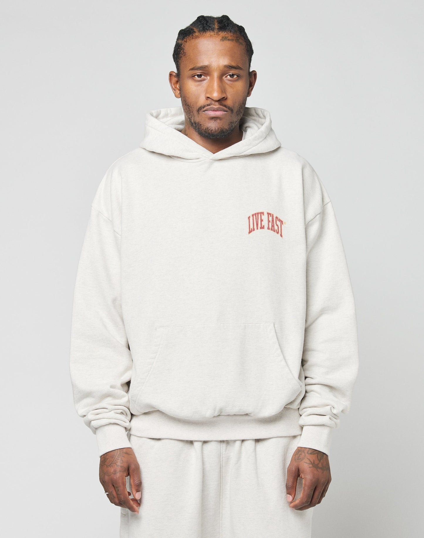 A man faces forward in a light beige 100% Baumwolle Live Fast Amsterdam Hooded by LIVE FAST DIE YOUNG, paired with matching sweatpants. An oversize fit, it sports LIVE FAST in red on the chest. The background is plain and light-colored.