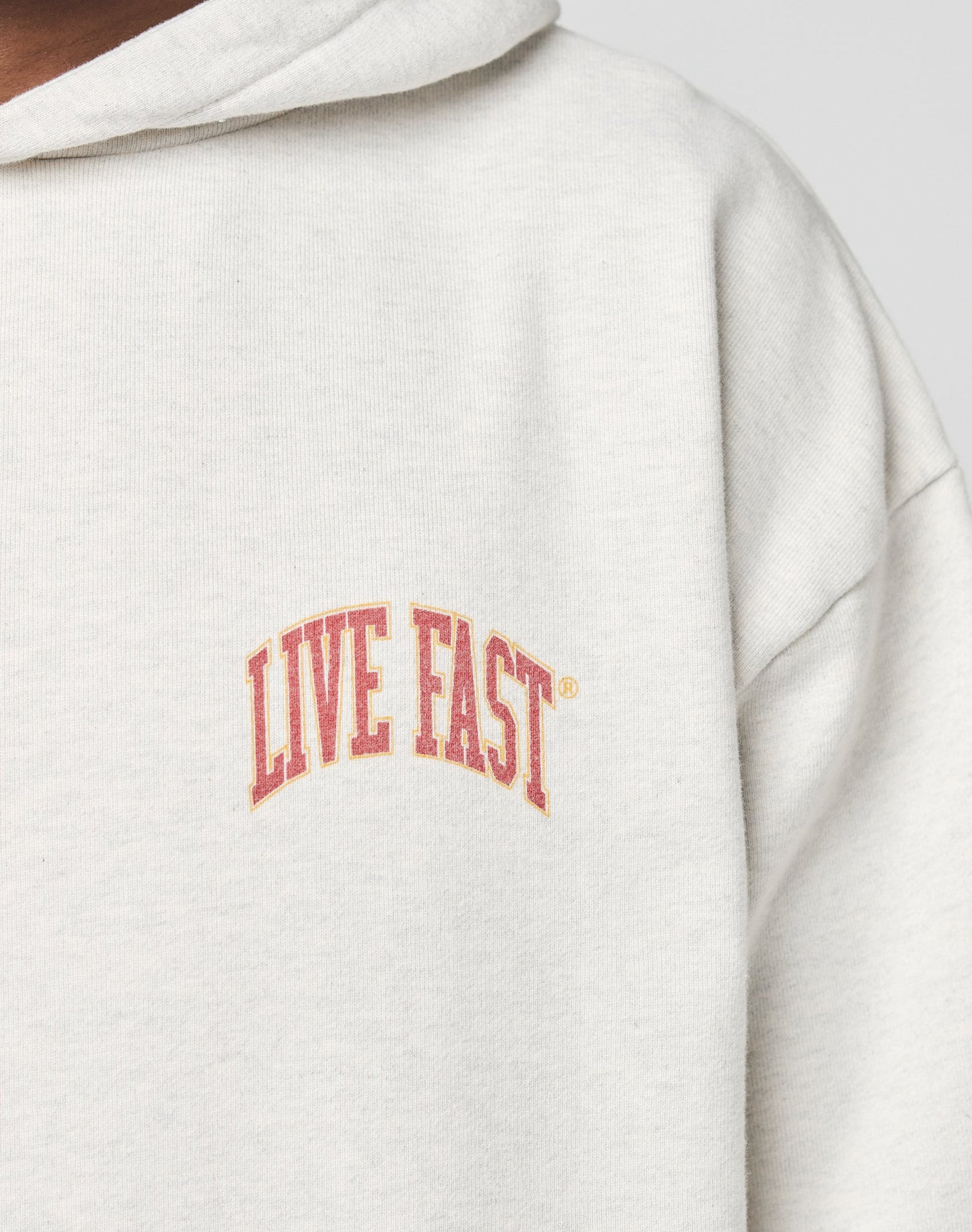 A close-up of the Live Fast Amsterdam Hooded by LIVE FAST DIE YOUNG, an oversize light gray hoodie with bold, red arched LIVE FAST text on the chest. Made from soft 100% Baumwolle; hood partially visible.