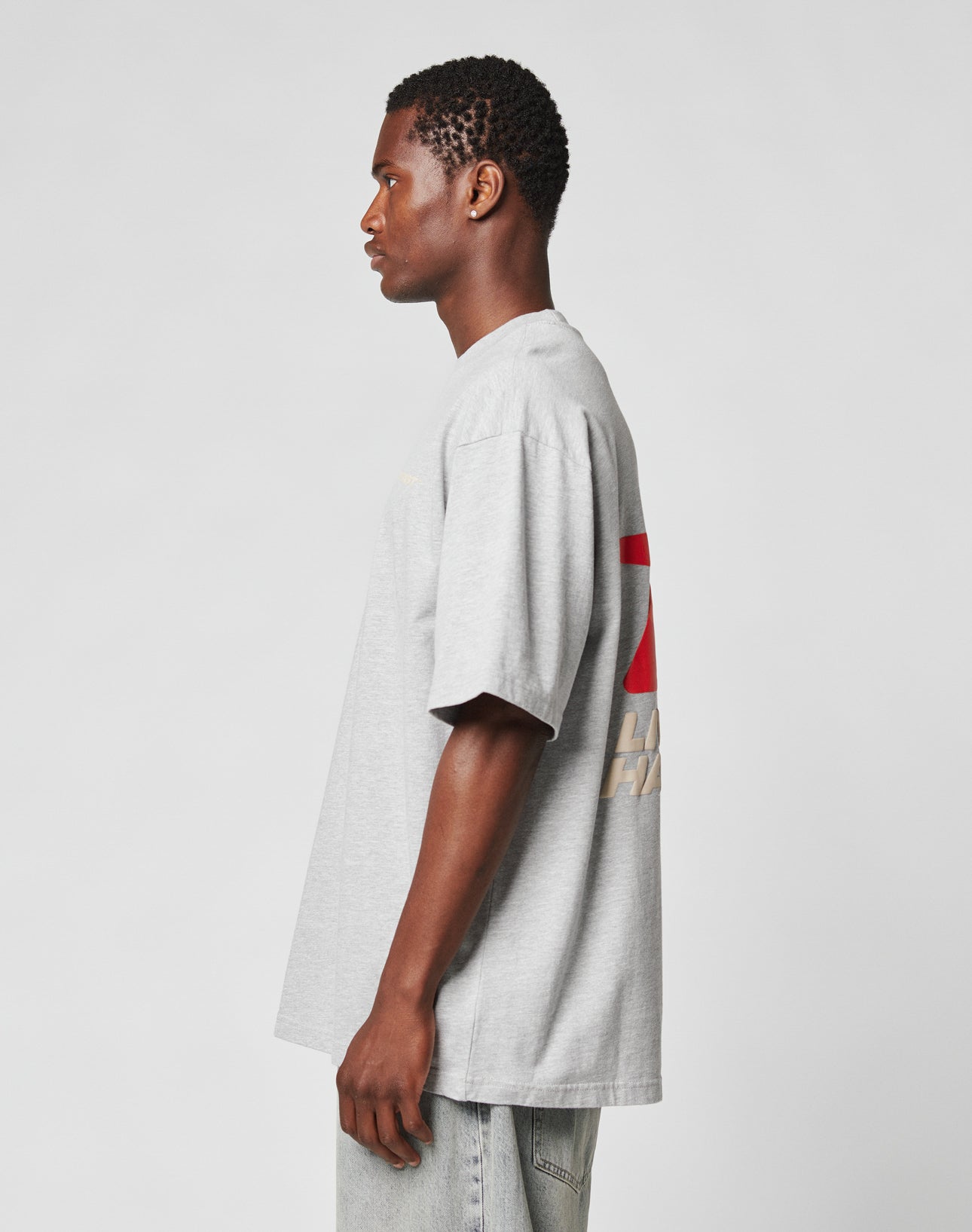 Someone in profile wears the oversize LFDY Hardwear Tee by LIVE FAST DIE YOUNG in light gray, featuring a red and white back design, paired with light blue jeans against a plain light background.