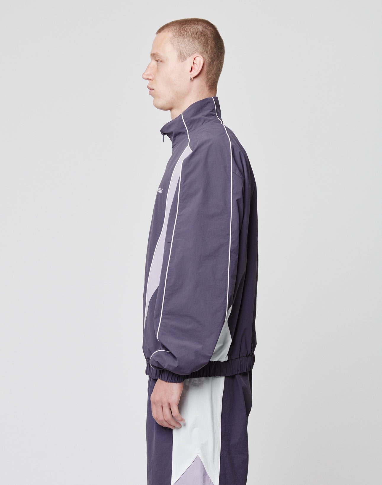 A man with short hair stands in profile, wearing the LIVE FAST DIE YOUNG Basic Nylon Trackjacket in purple and white with a high collar, white stripes, and side pockets, set against a plain light gray background.
