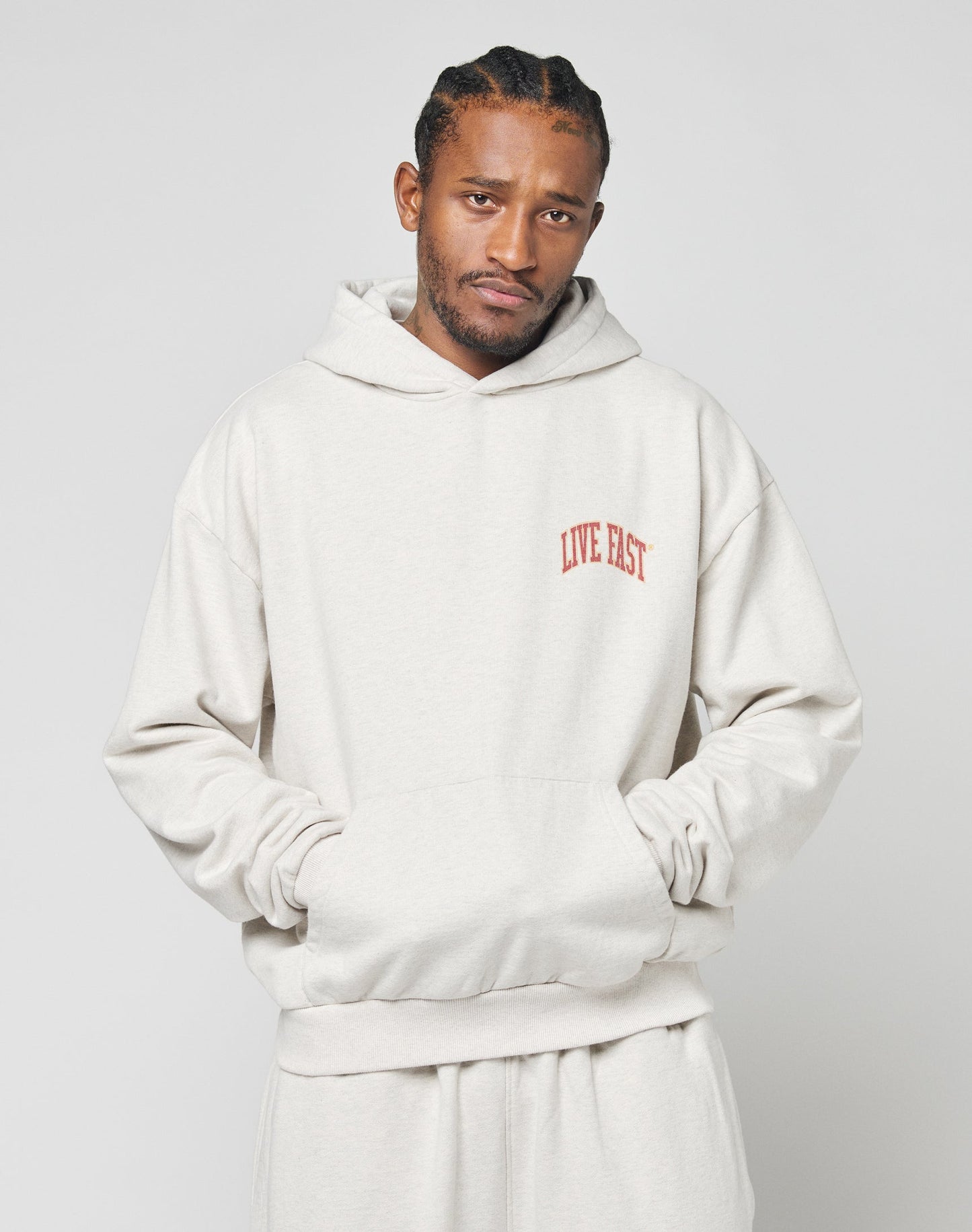 A man with braided hair wears the Live Fast Amsterdam Hooded sweatshirt and matching pants by LIVE FAST DIE YOUNG, in oversize fit light beige 100% Baumwolle, featuring a red LIVE FAST chest print. He stands hands in pockets against a plain background.