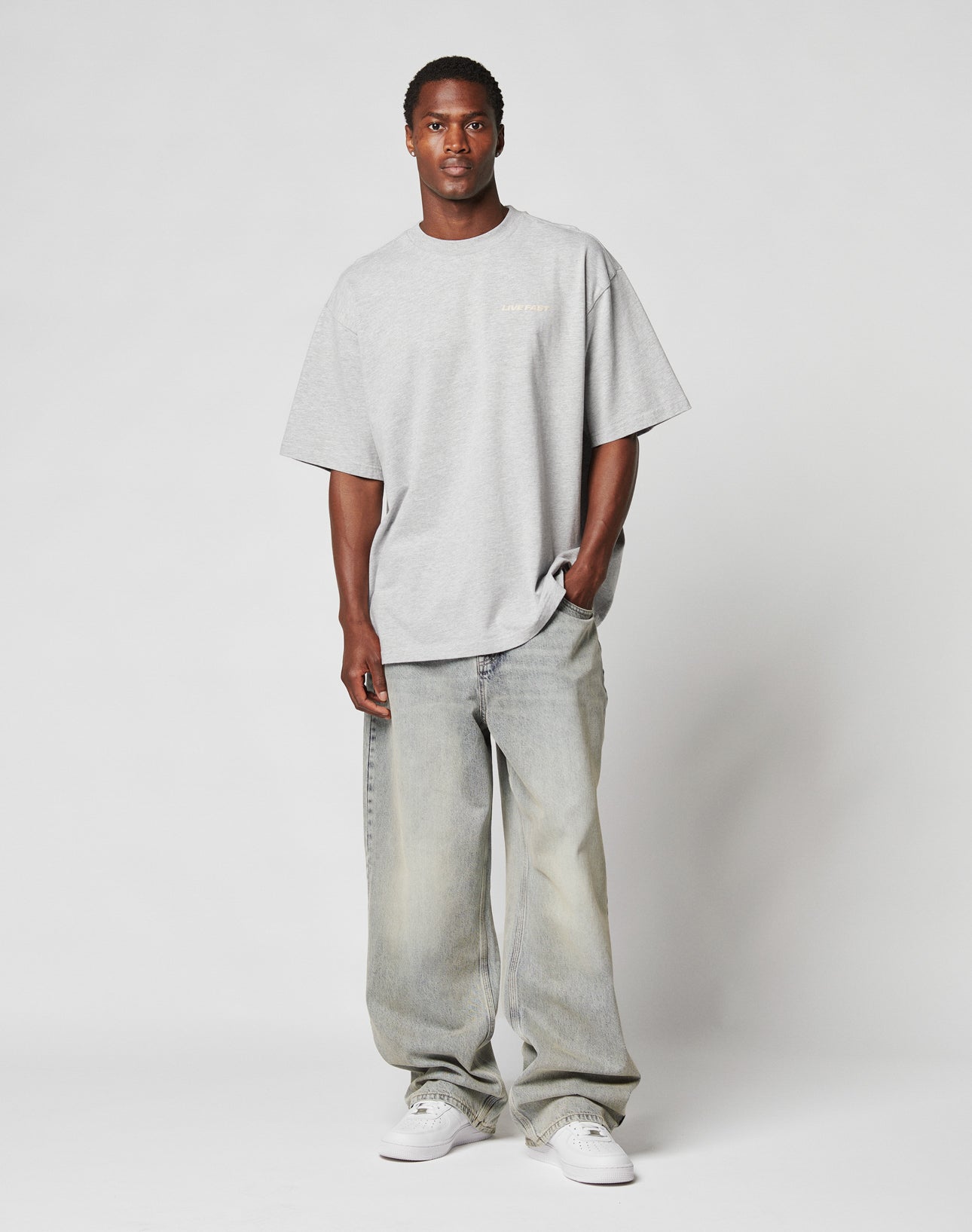 A man stands against a plain white background wearing the LIVE FAST DIE YOUNG LFDY Hardwear Tee, baggy faded jeans, and white sneakers. One hand is in his pocket as he looks straight ahead with a neutral expression.