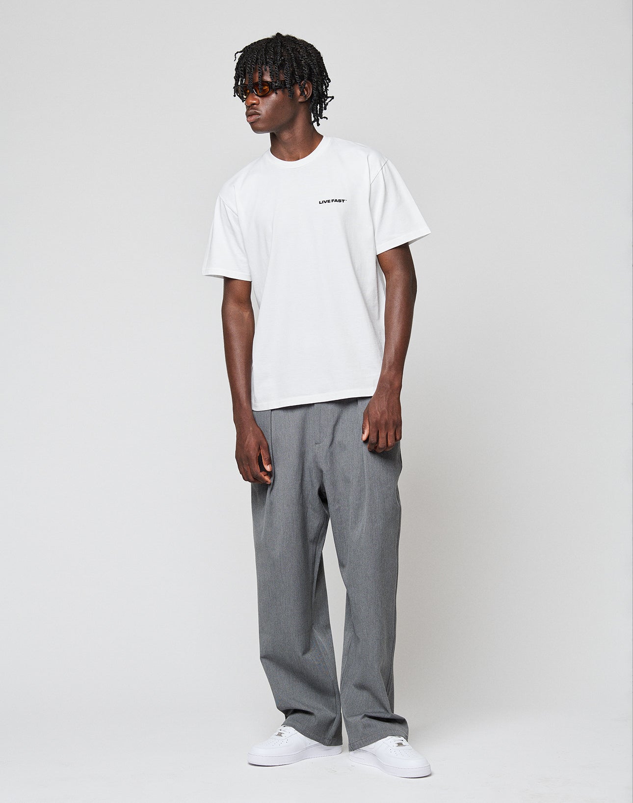 A person with short, textured hair stands against a plain background, wearing the LIVE FAST DIE YOUNG Live Fast Plain Tee, loose gray pants, and white sneakers. They have a relaxed posture and a neutral expression.