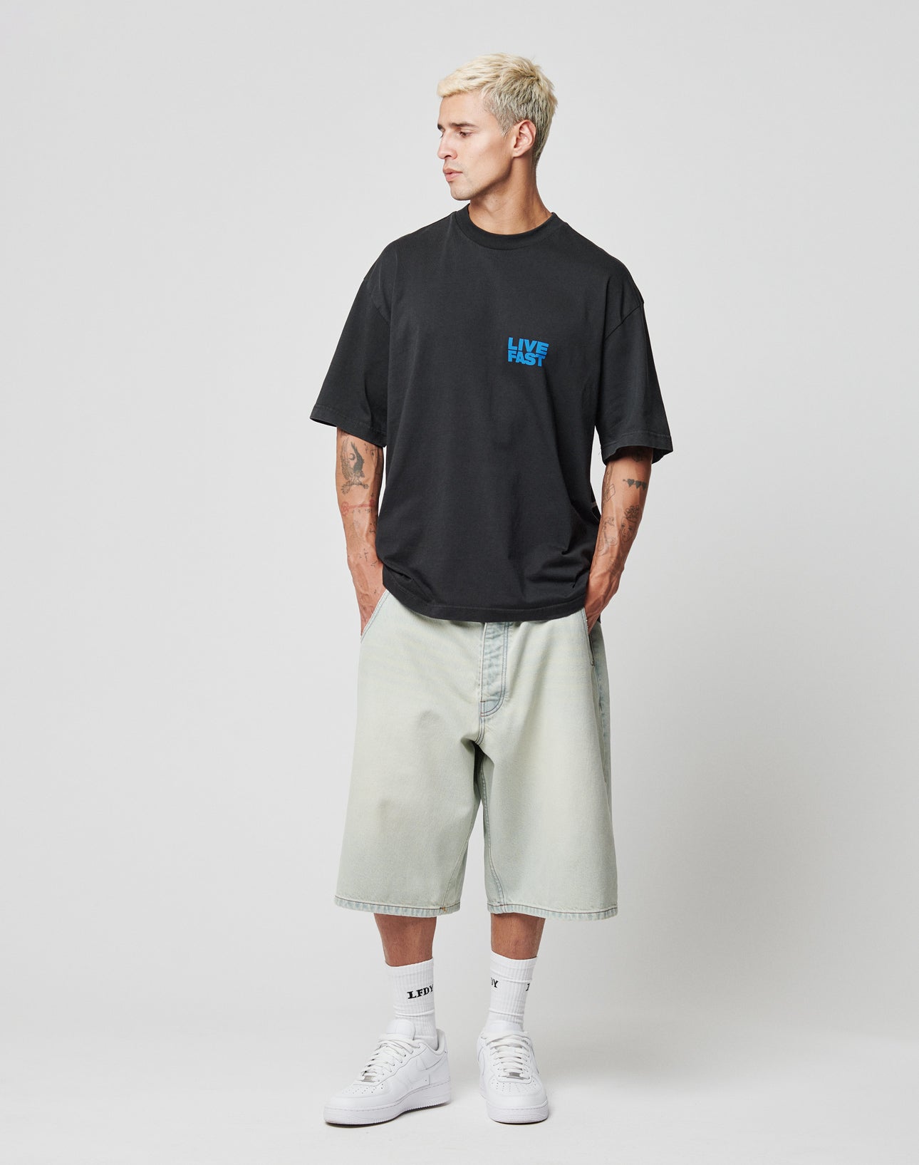 A man with short blond hair wears a loose black T-shirt, LIVE FAST DIE YOUNG LFDY Jorts with Gesäßtasche, white socks, and white sneakers. He stands hands in pockets, looking to the side against a plain white background.