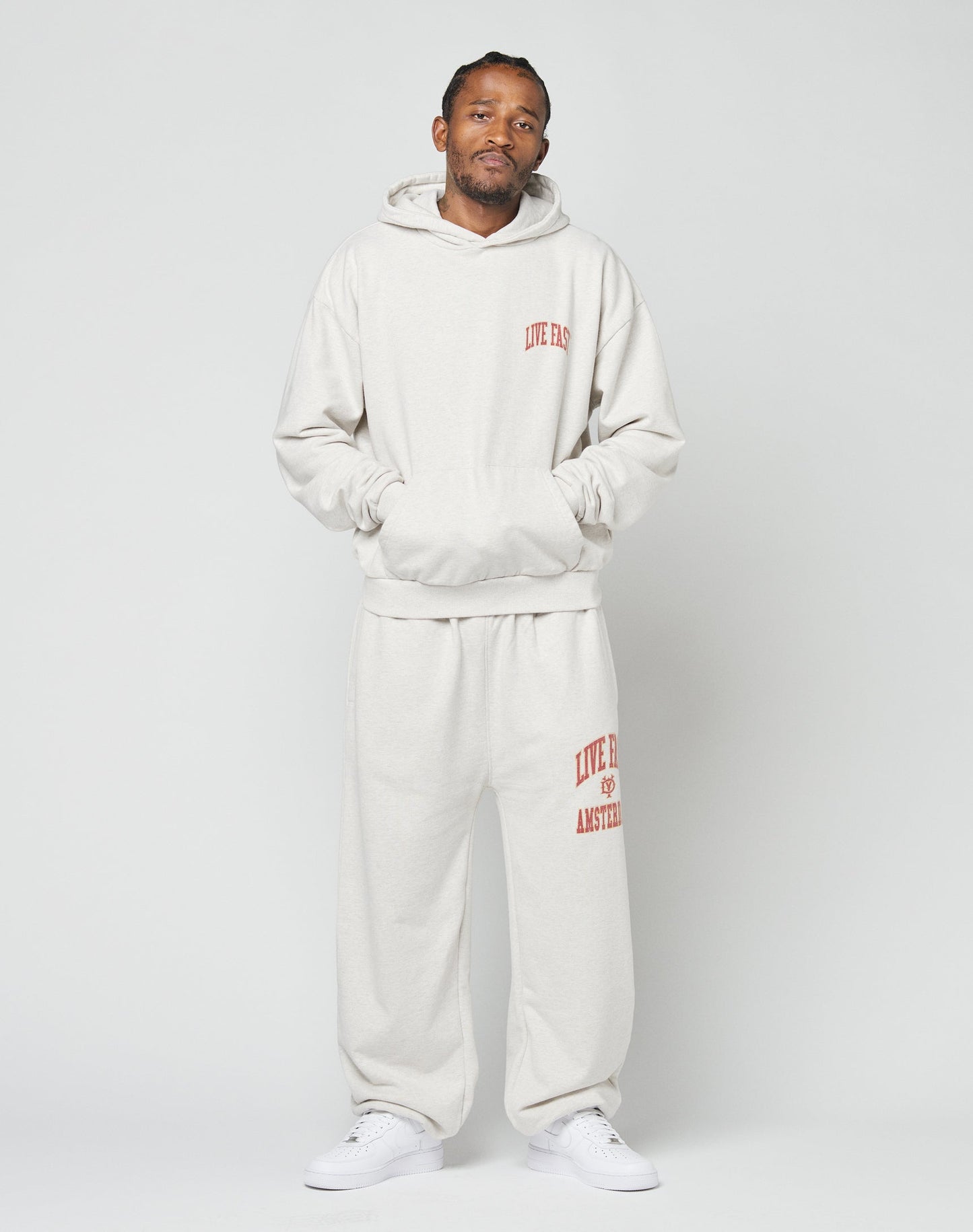 A man stands against a plain background wearing the LIVE FAST DIE YOUNG Live Fast Amsterdam Loose Jogger and matching light gray hooded sweatshirt with red text, paired with white sneakers. His hands are in the sweatshirt pocket as he looks at the camera.