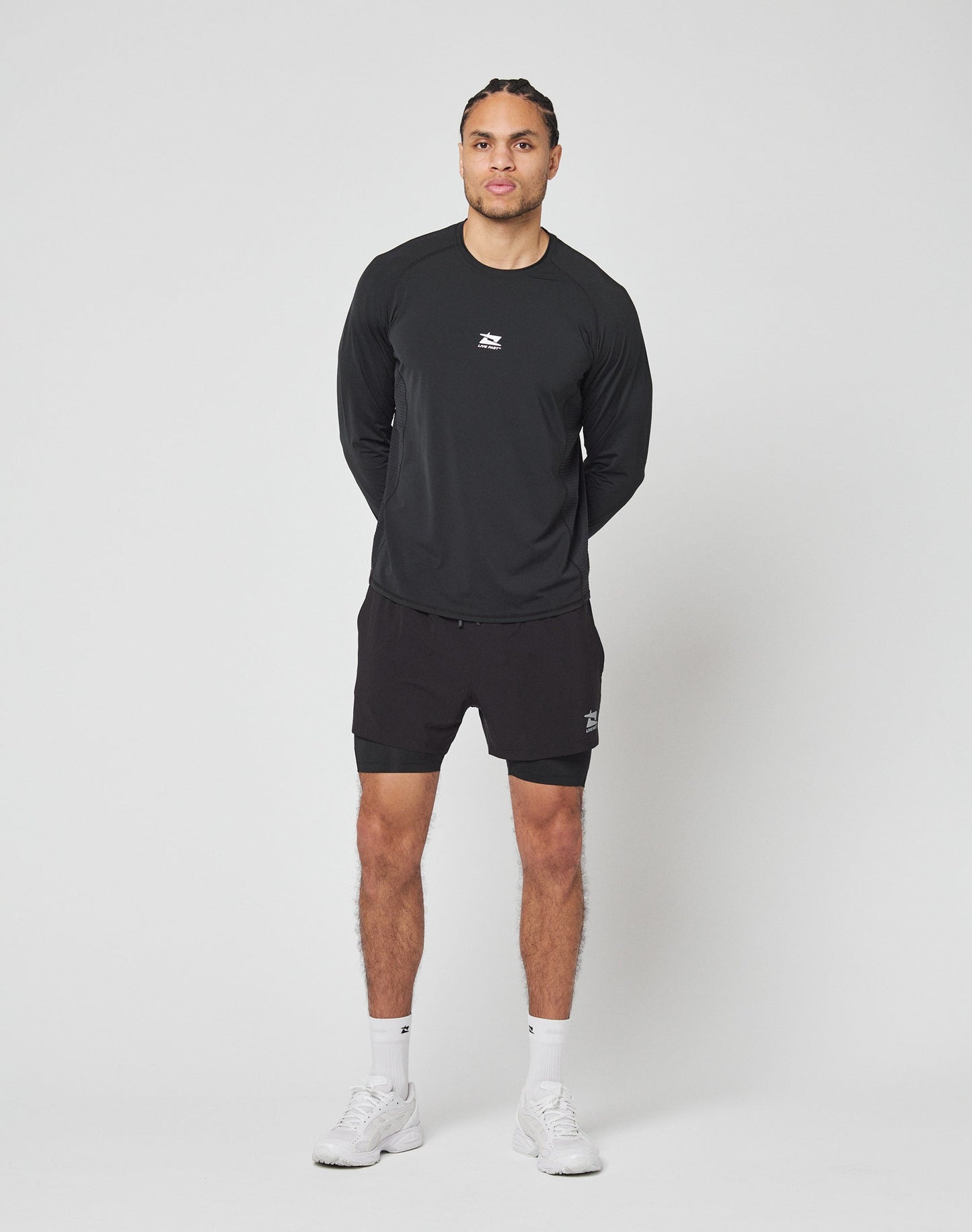 A person stands against a plain white background, wearing LIVE FAST DIE YOUNGs black Live Fast Performance Running Shorts with integrated tights, a black long-sleeve athletic shirt, white socks, and white sneakers. Hands behind back, facing forward.