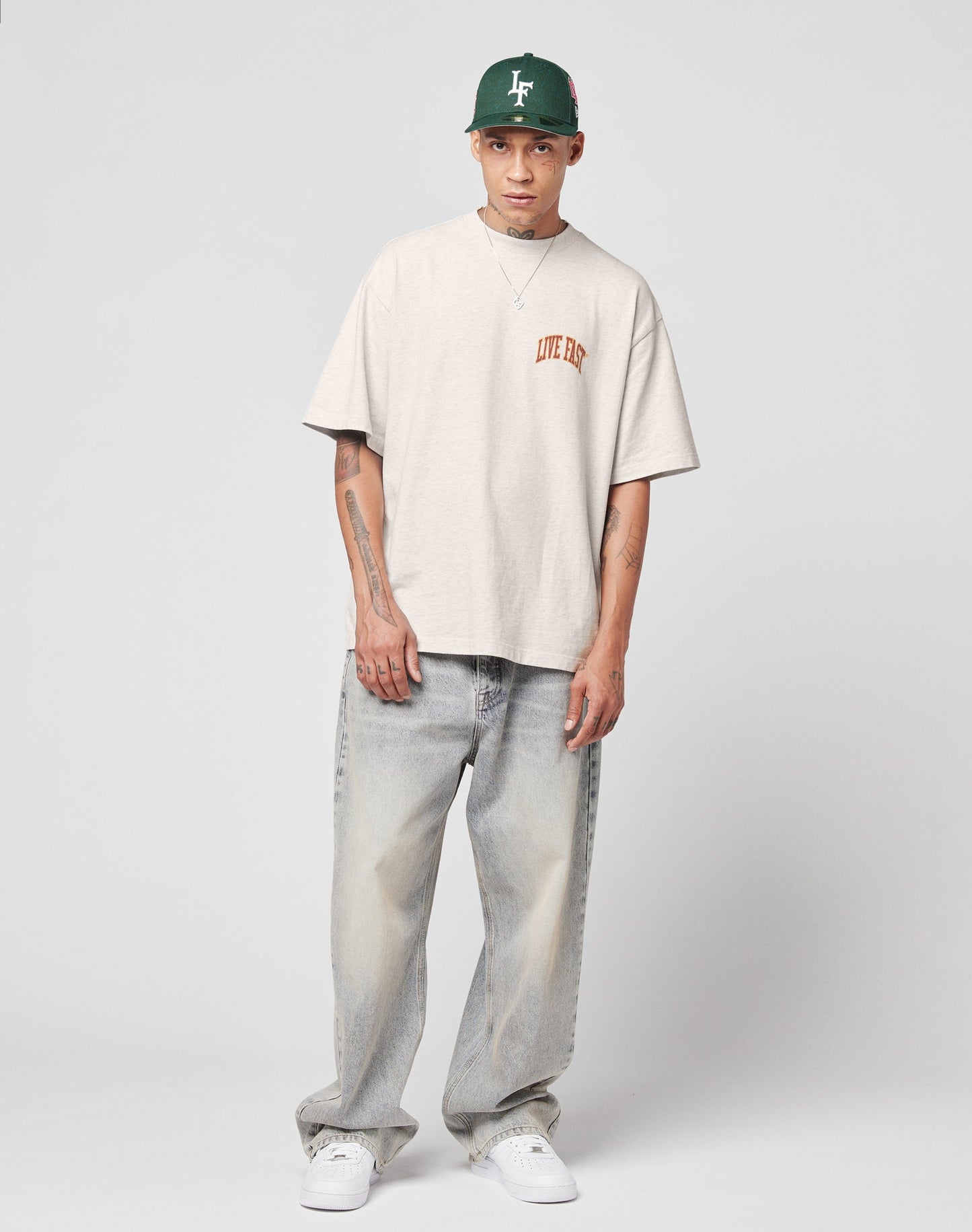 A person in a green cap, beige oversized Live Fast T-shirt, and white sneakers wears LIVE FAST DIE YOUNGs LFDY Baggy Denim—light blue, baggy fit jeans with wide straight legs and a subtle flag label—against a plain gray background.