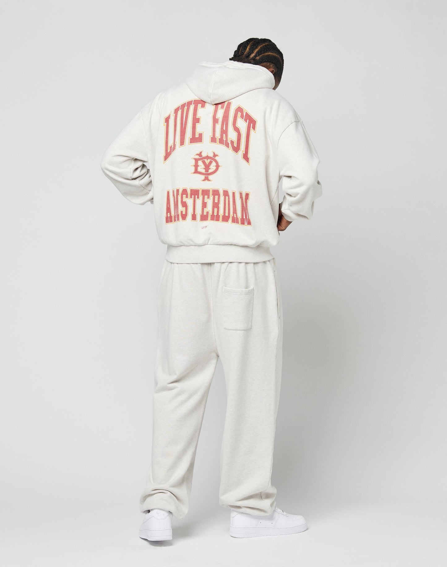Wearing the Live Fast Amsterdam Hooded by LIVE FAST DIE YOUNG, a person in an off-white, oversized hoodie and sweatpants faces away, showing a bold backprint. Made from 100% Baumwolle and paired with white sneakers.