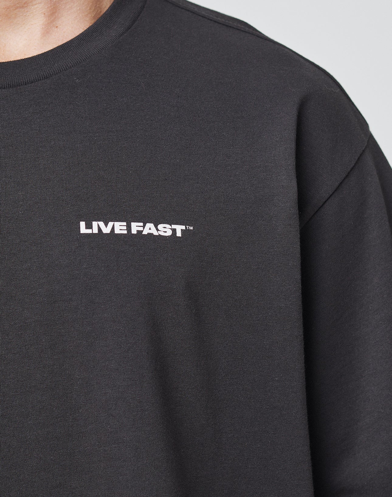 A close-up of a person in the LIVE FAST DIE YOUNG LFDY Cinema Tee, featuring LIVE FAST™ printed in white on the left chest. Only part of the neck, shoulder, and upper chest are visible. Oversize fit for a relaxed look.